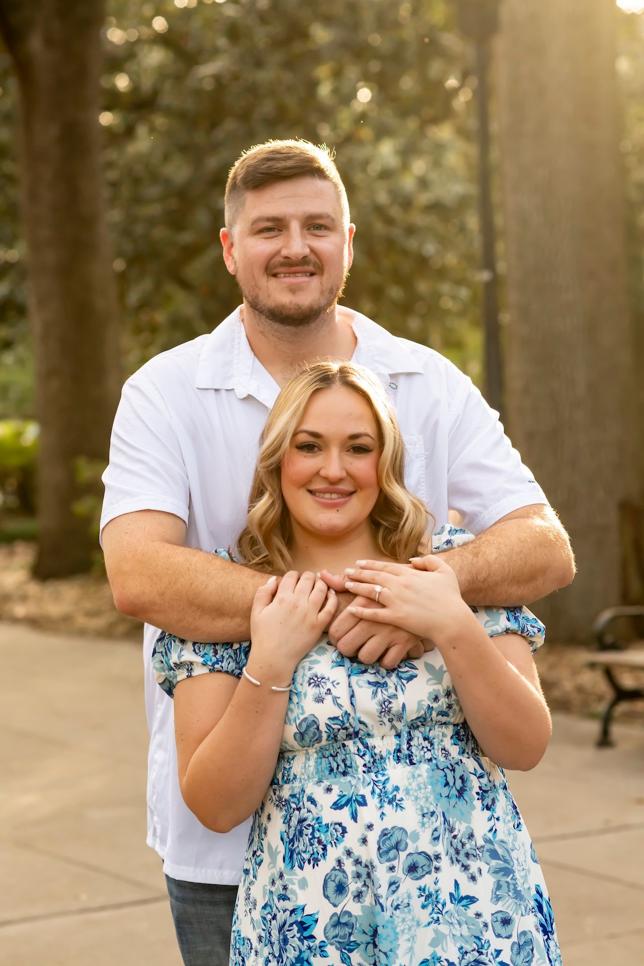 Couple embraces outdoors in a romantic pose during golden hour, wearing white and blue floral clothing.