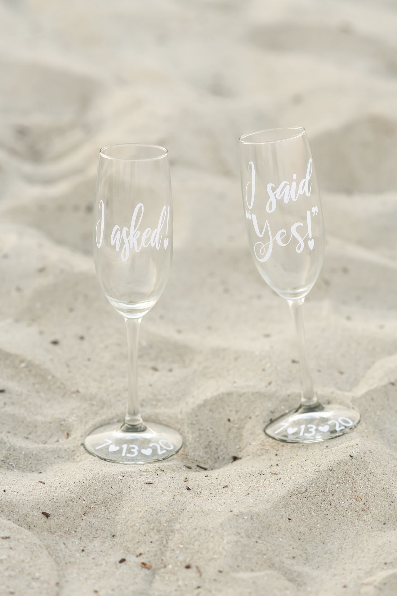 Two champagne flutes in sand with 'I asked' and 'Yes!' written in white script lettering.