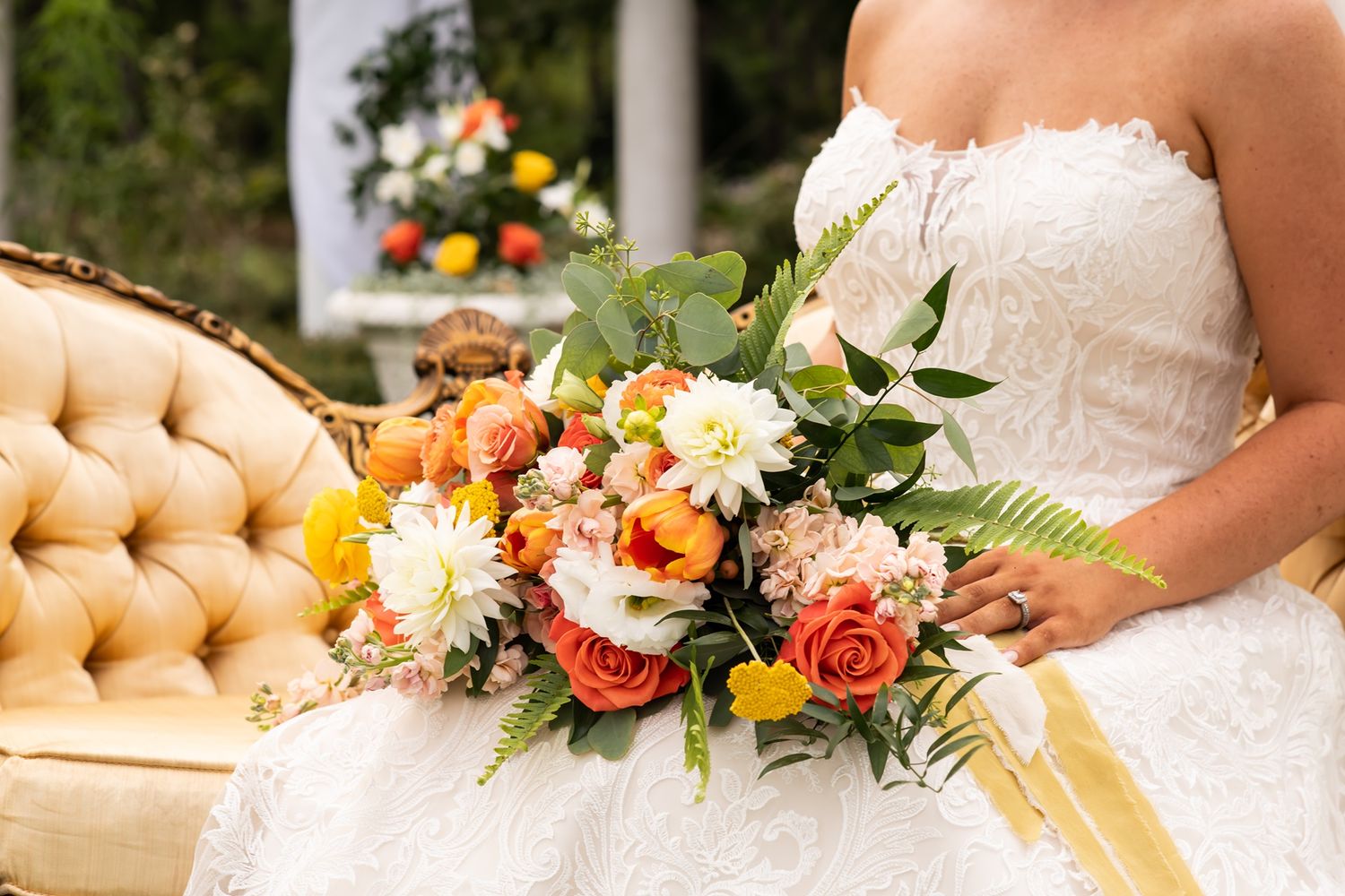 Vibrant bridal bouquet with orange and peach roses rests on tufted champagne velvet couch.