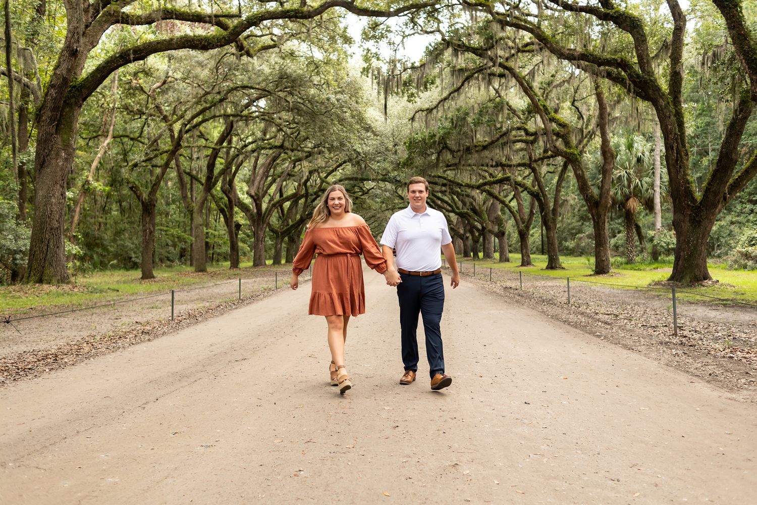 A couple walks hand in hand down a tree-lined dirt path in a rustic outdoor setting.
