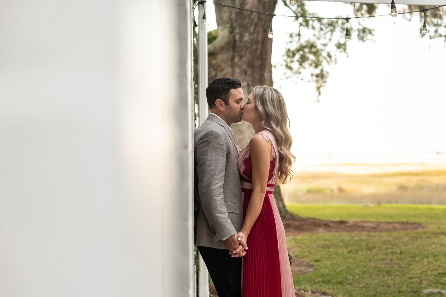 Romantic couple shares tender moment while standing by water with natural scenery in background.