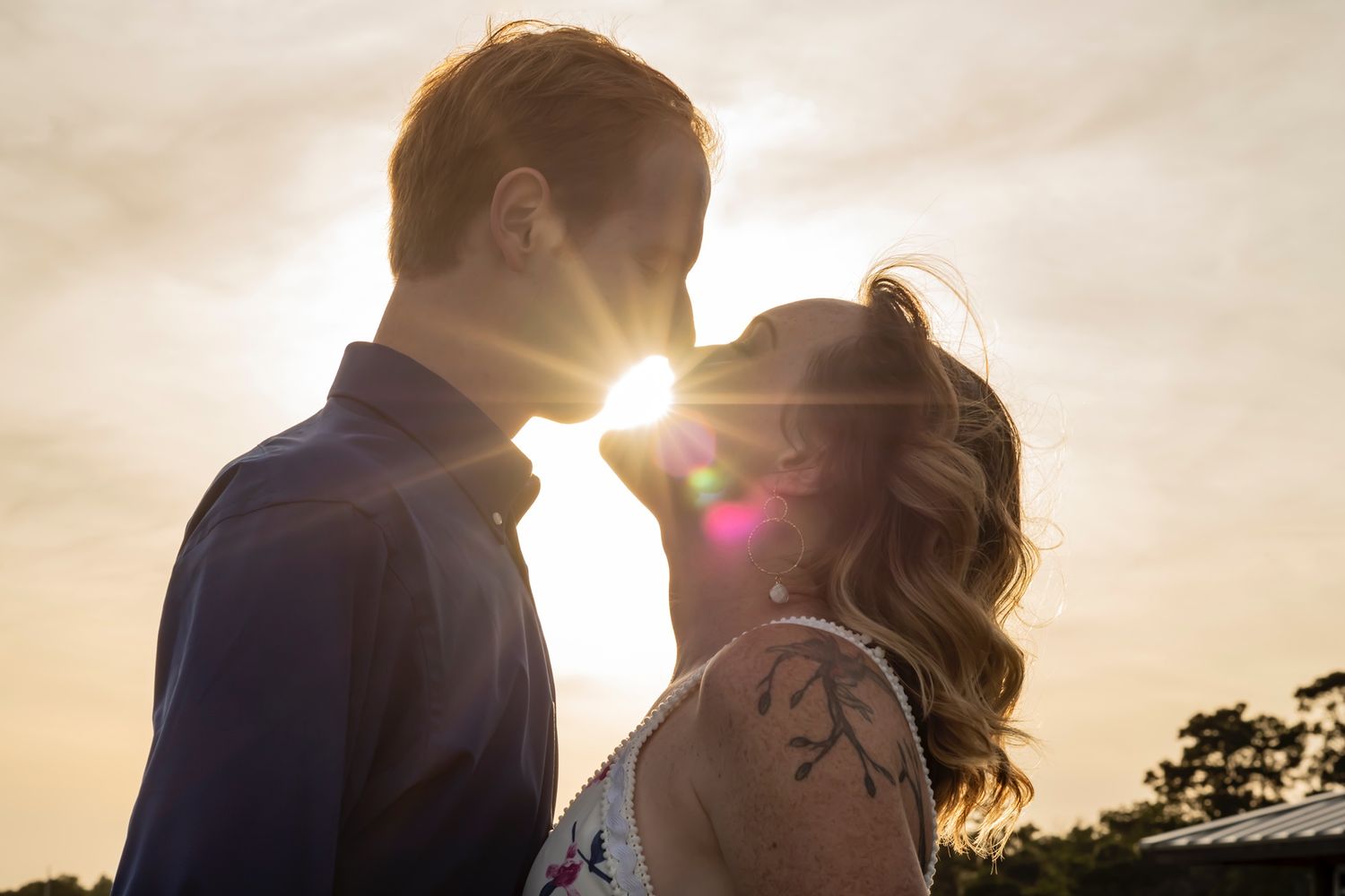 Two silhouettes stand close together against a bright sunset sky with lens flare and trees in the background.