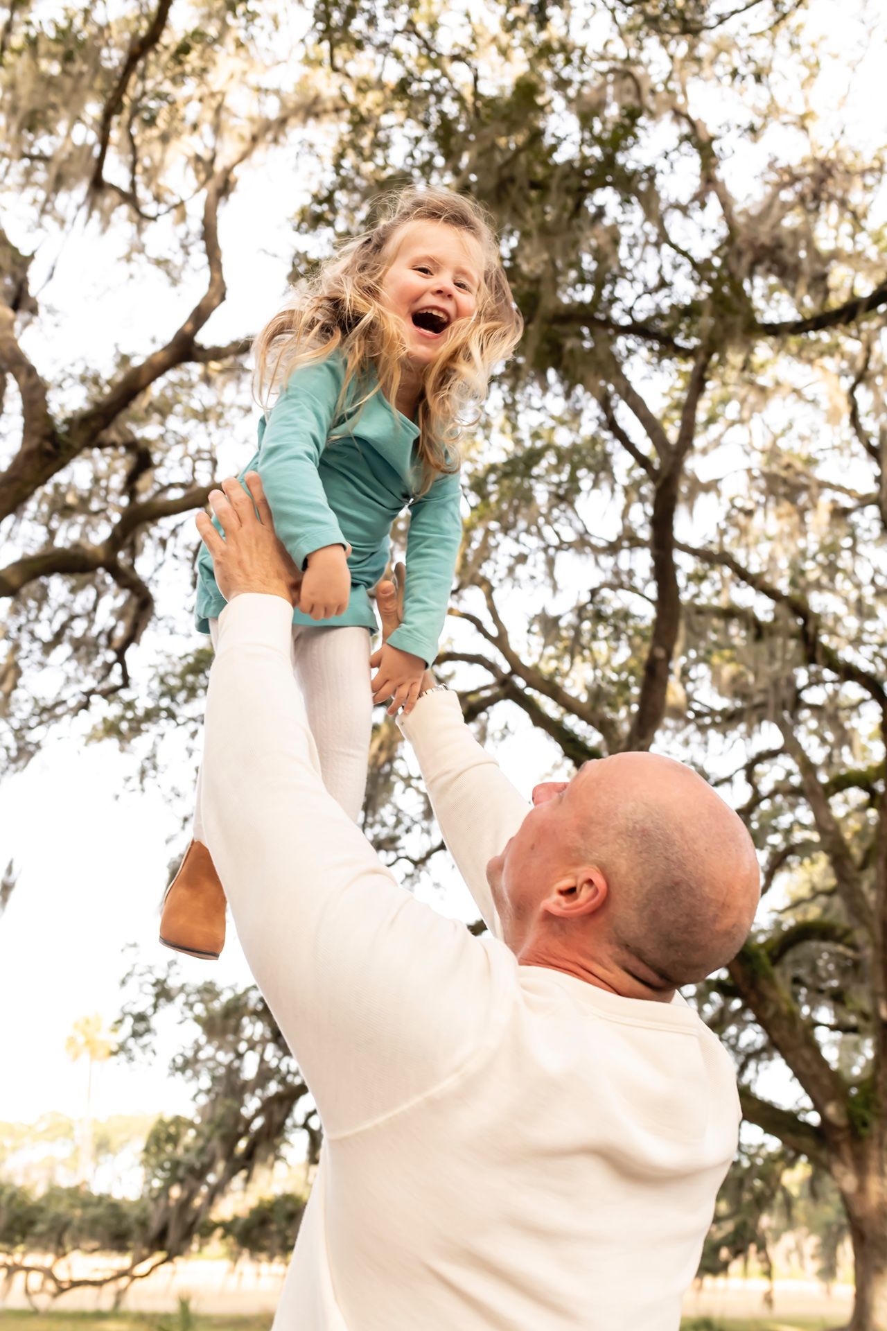 A person in a cream sweater lifts a laughing child wearing teal in front of mossy oak trees in an outdoor setting.