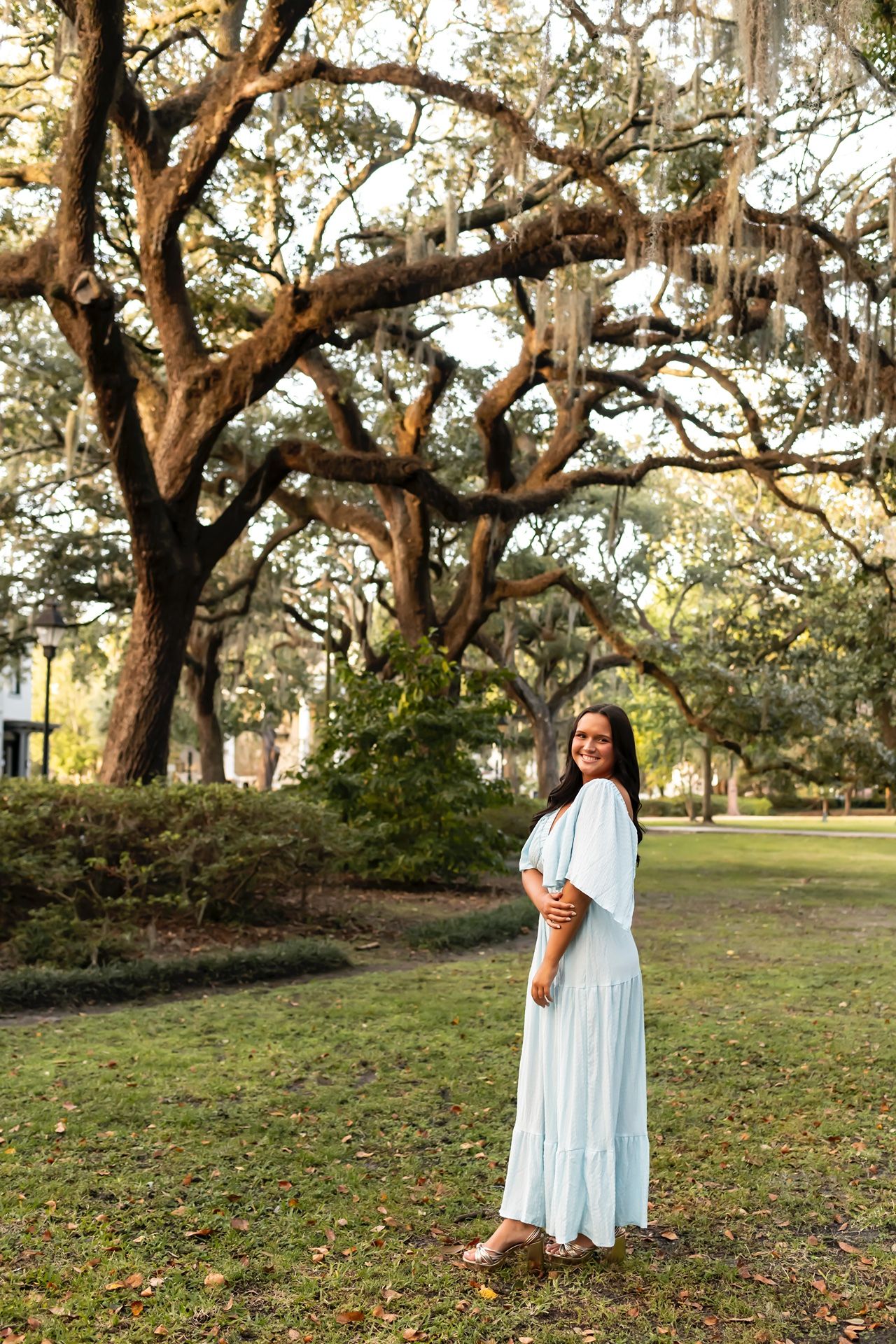 A person in a flowing white dress poses near a majestic Spanish moss-draped oak tree in a serene park setting.