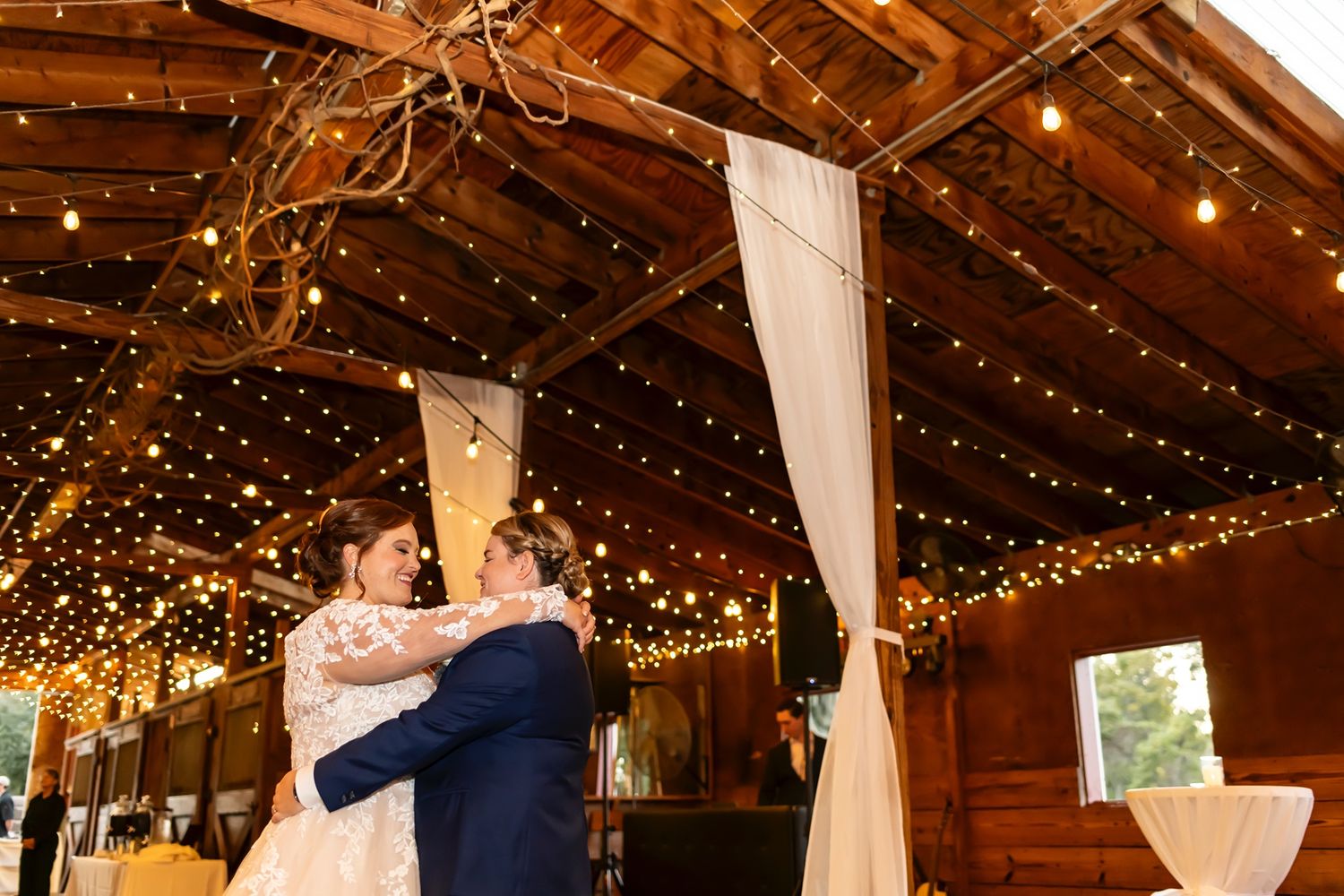 A couple shares a romantic dance under twinkling string lights in a rustic wooden barn reception venue.