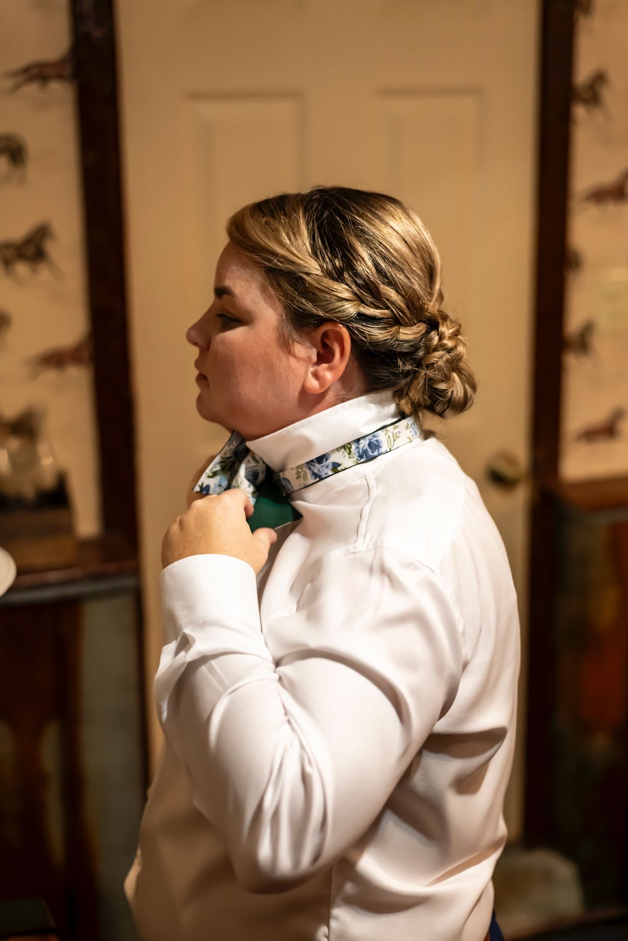 Someone wearing a white dress shirt adjusts their collar while preparing in a room with equestrian wall decor.