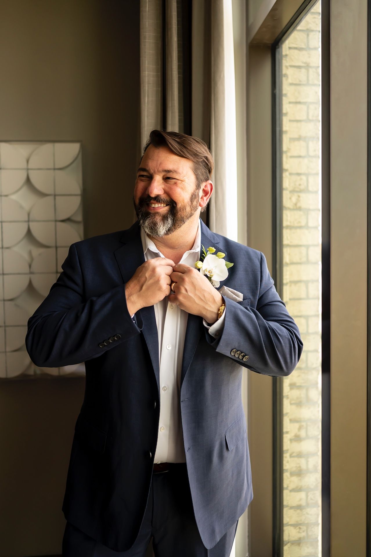 A groom in a navy suit adjusts his bow tie while standing by a window in natural light.