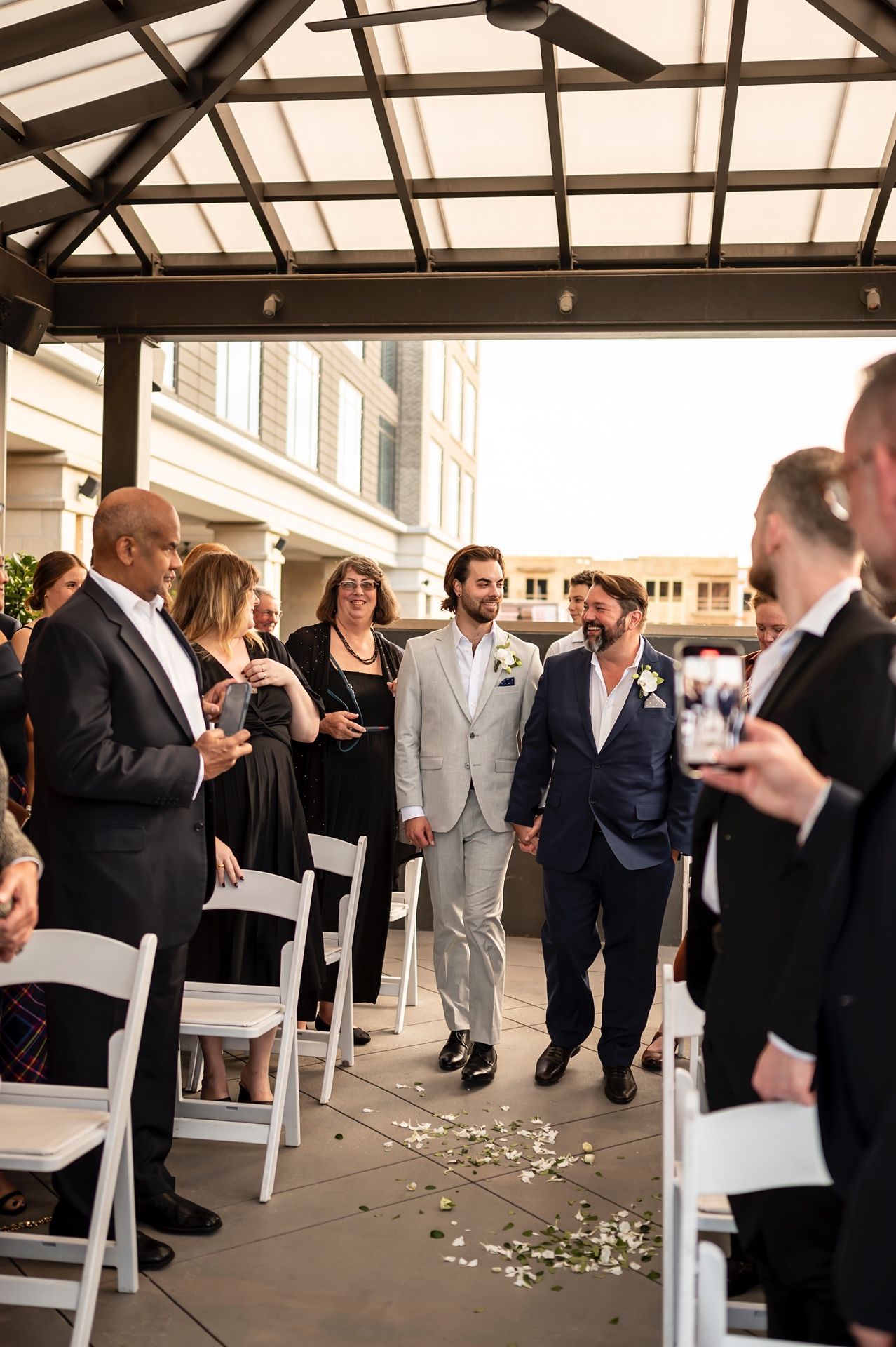 A group of wedding guests standing under a covered patio with white chairs and scattered flower petals on the ground.