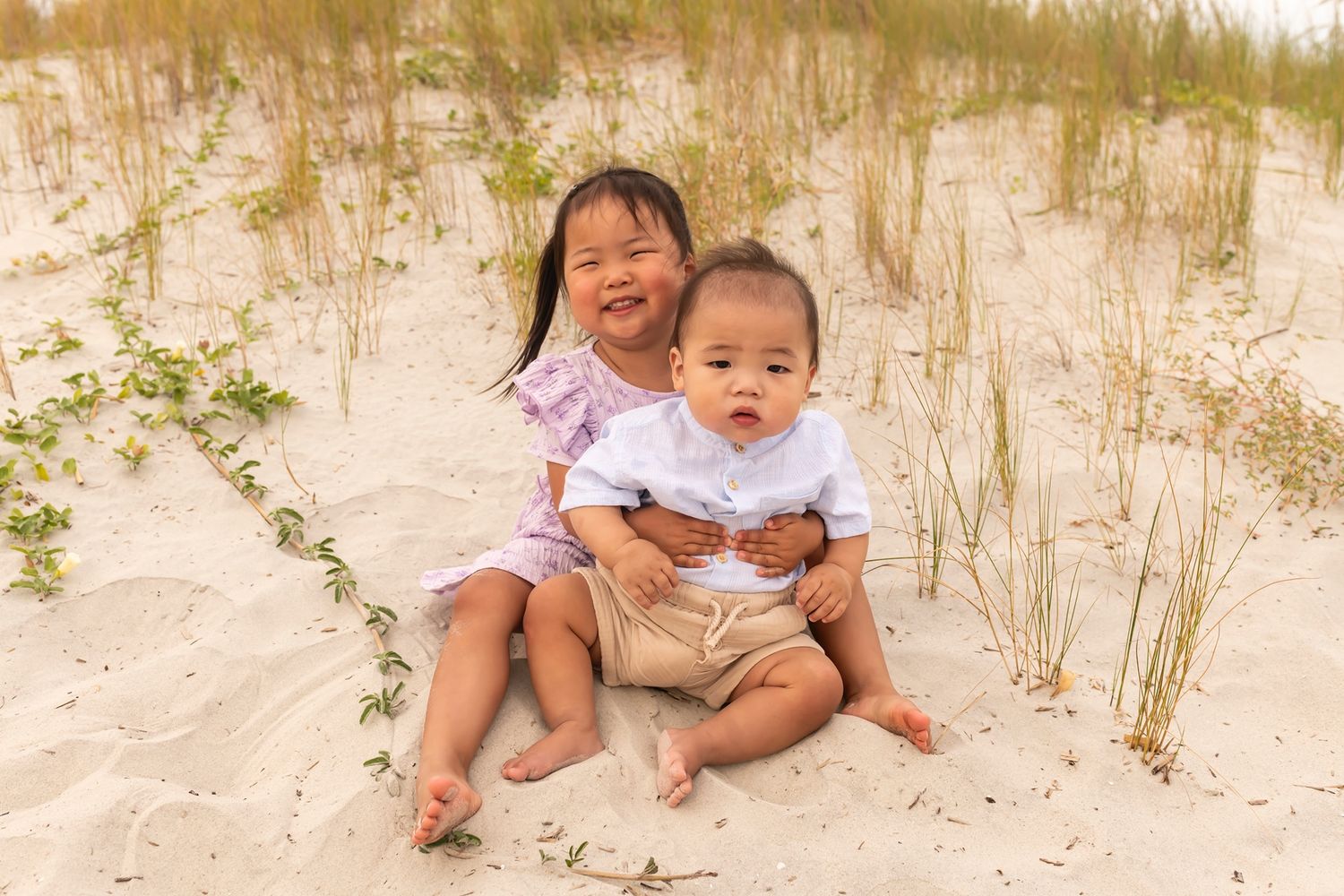 Two small children sitting together on sandy beach dunes with grass during golden hour.
