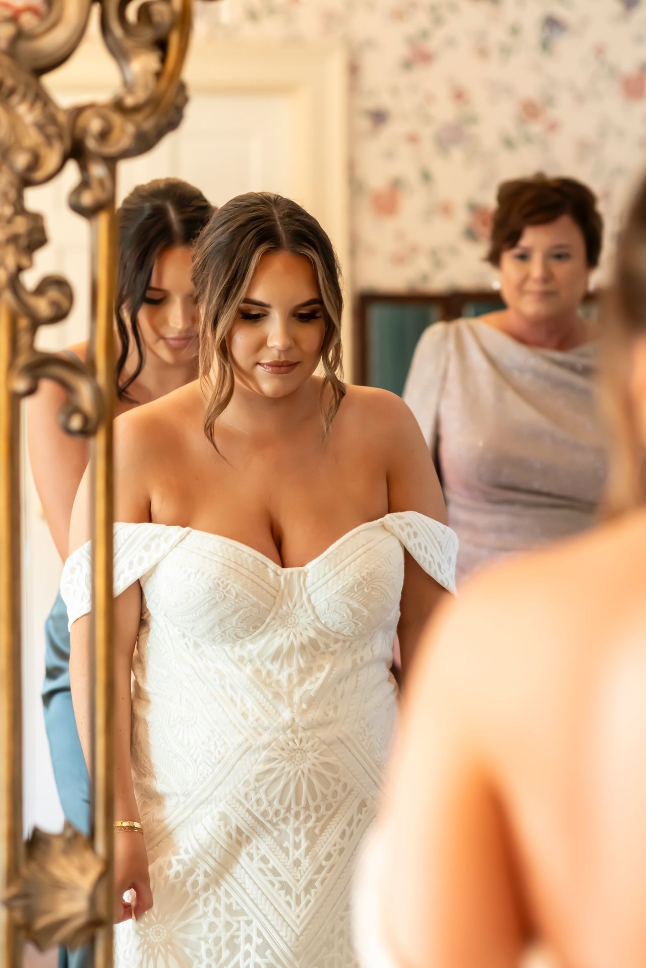 Bride wearing an off-shoulder white geometric patterned wedding dress looks at herself in an ornate gold mirror.