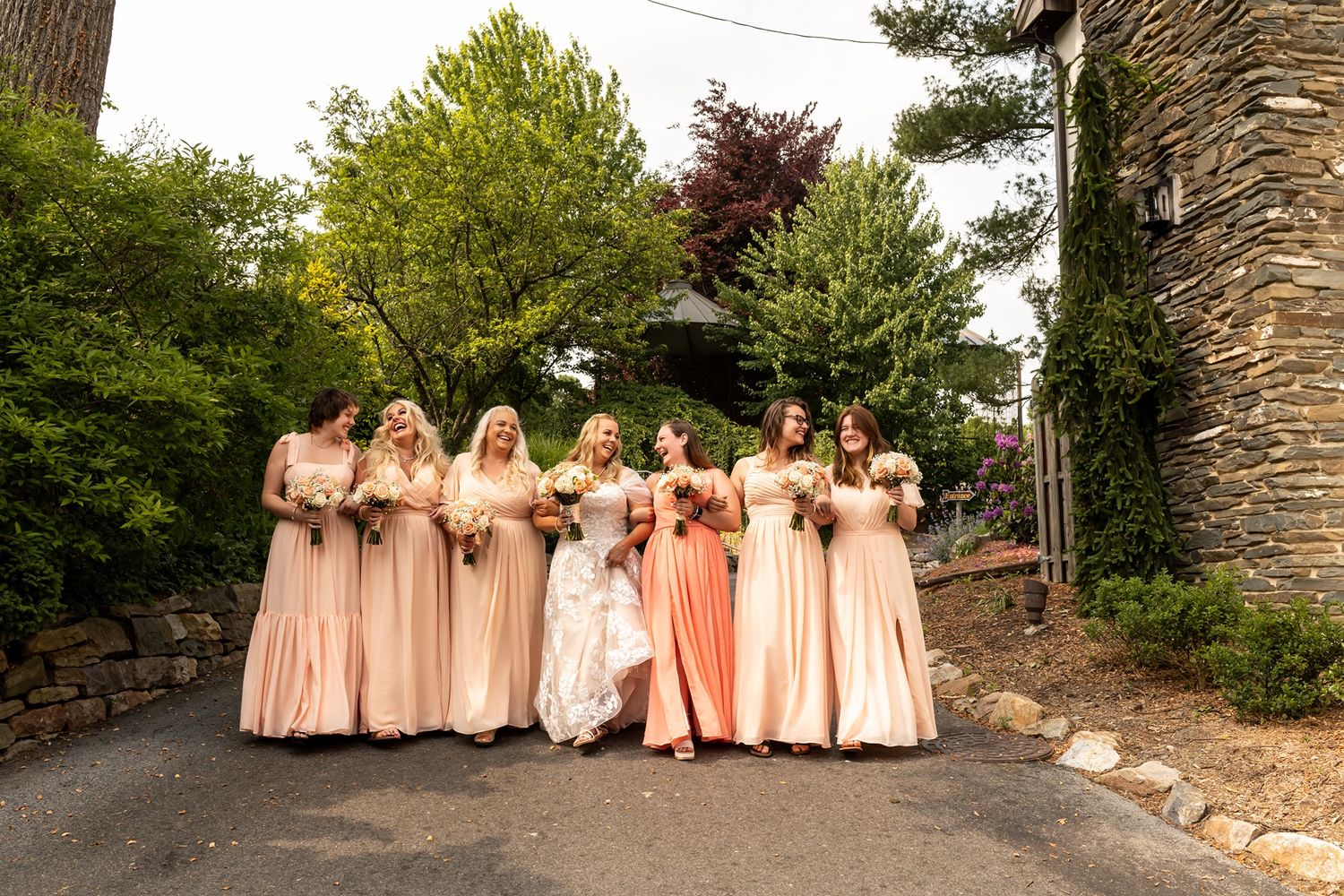 Bridesmaids in flowing peach dresses stand in line along a stone path surrounded by green foliage.