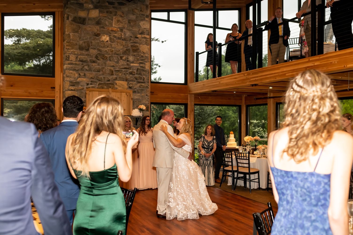 Wedding couple sharing a dance surrounded by guests in a rustic venue with large windows and stone walls.