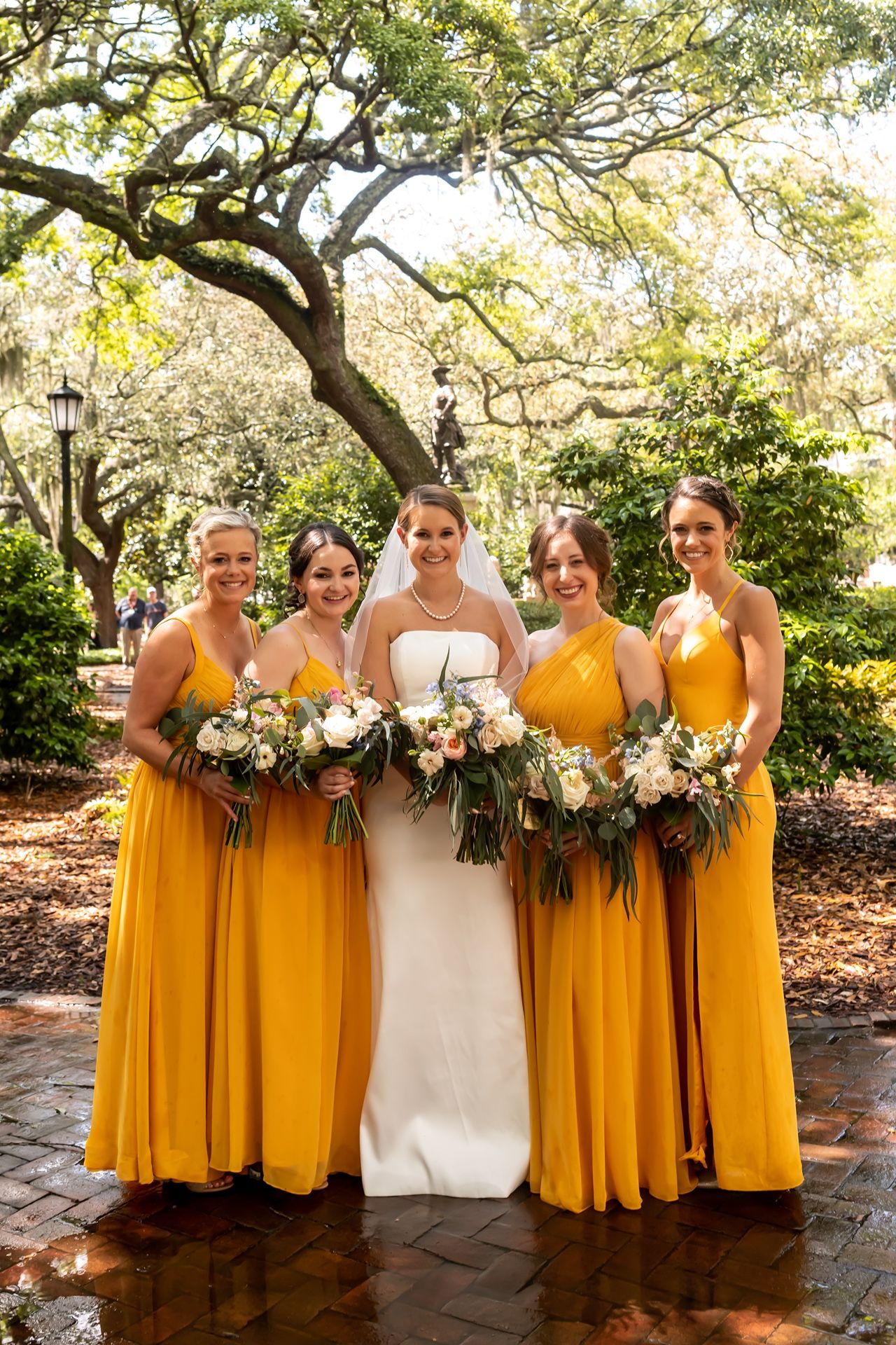 Wedding party wearing marigold yellow bridesmaid dresses and white bridal gown pose together in a garden setting.