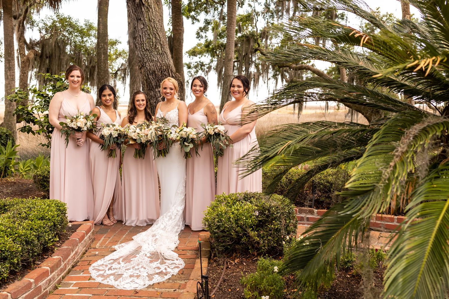 A bridal party in blush pink dresses poses on brick steps surrounded by tropical palm trees and lush gardens.