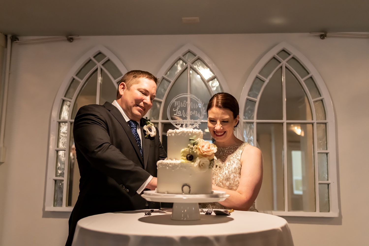 A happy couple cuts their elegant white wedding cake together during their reception while guests watch with joy.