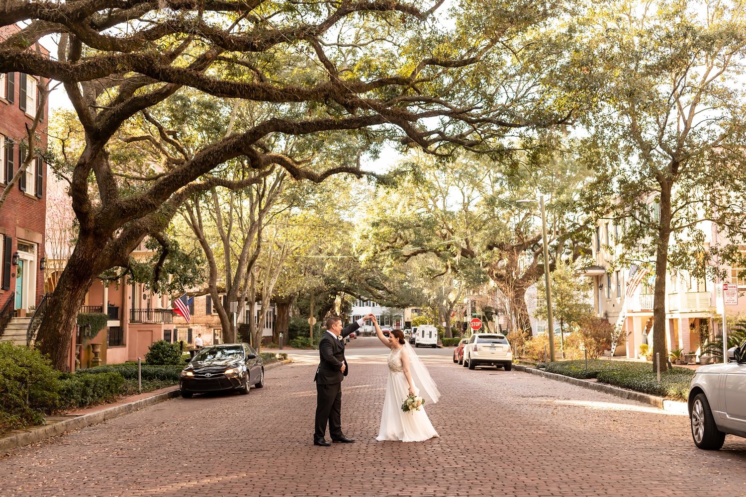 A couple stands together on a picturesque brick-paved street lined with blooming oak trees and parked cars in golden light.