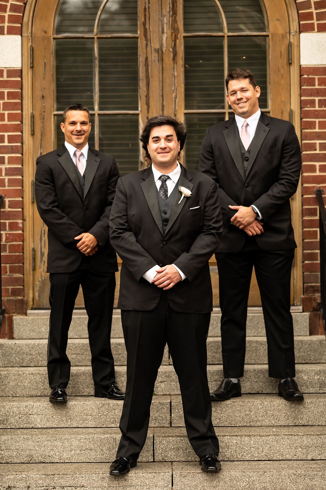 Three groomsmen in matching black suits and ties standing on brick steps in front of an arched wooden door.