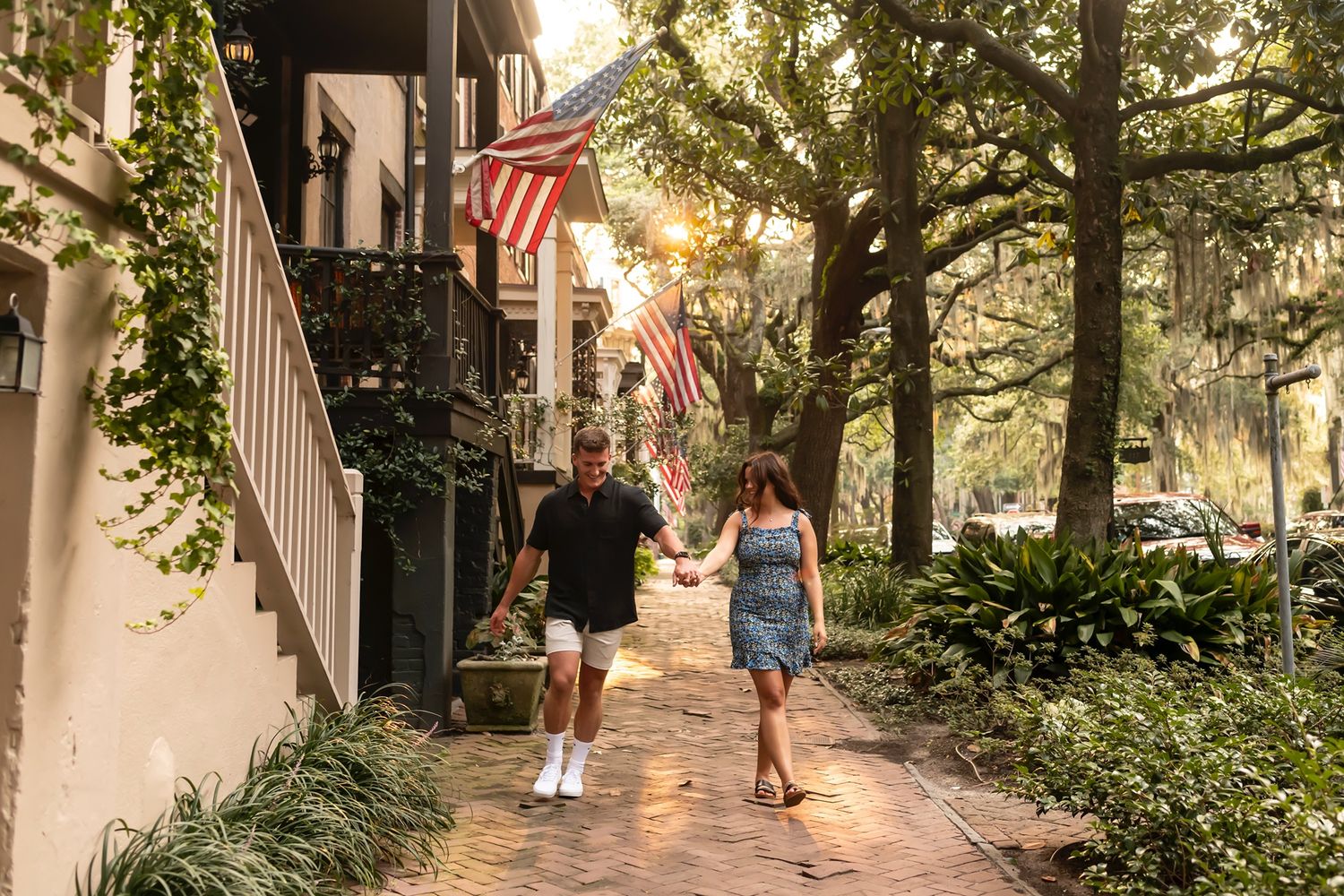 People walking on a charming cobblestone sidewalk with American flags and lush greenery in historic Savannah, Georgia at sunset.