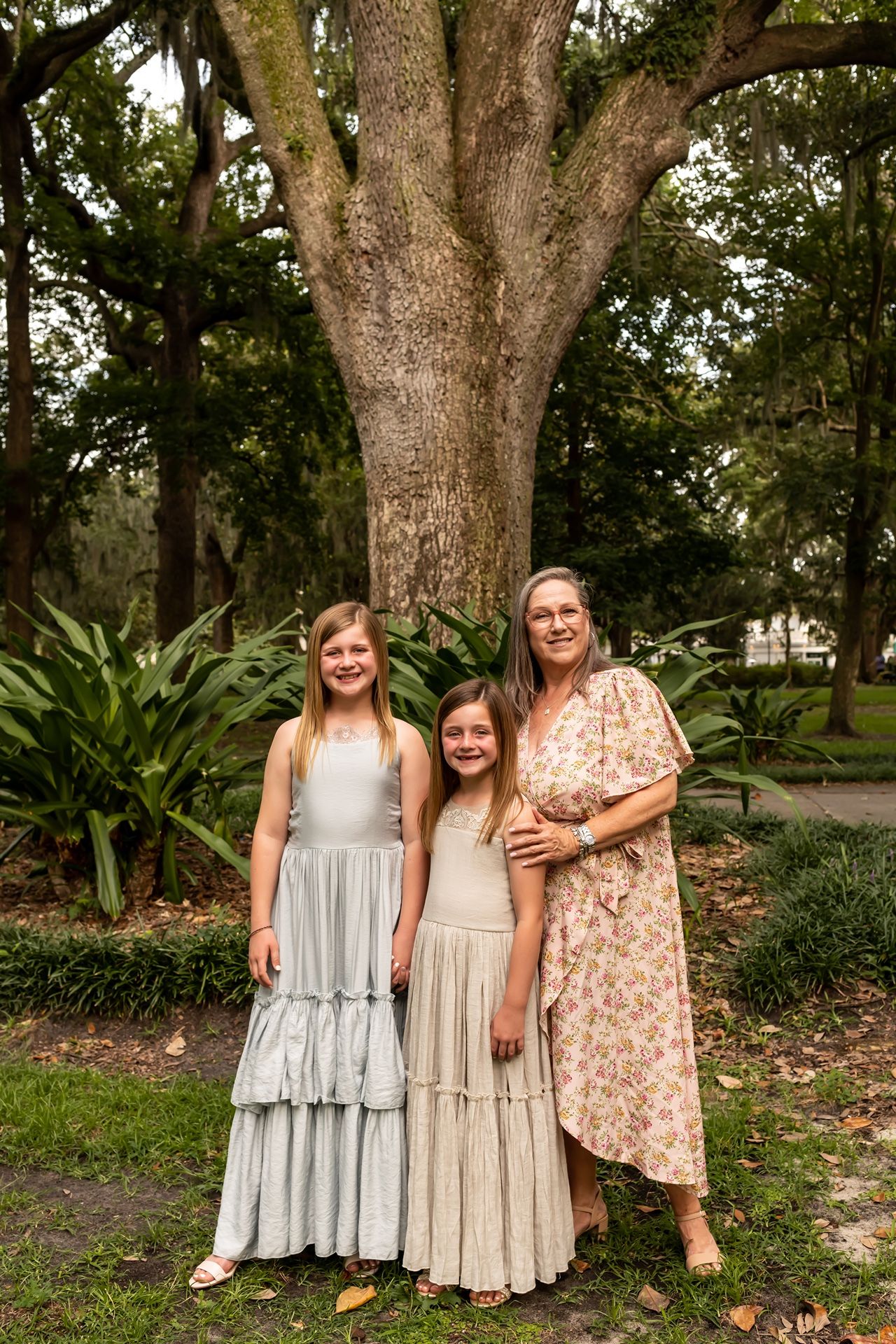 Three people in flowing dresses pose together in front of a large oak tree in a scenic garden setting.