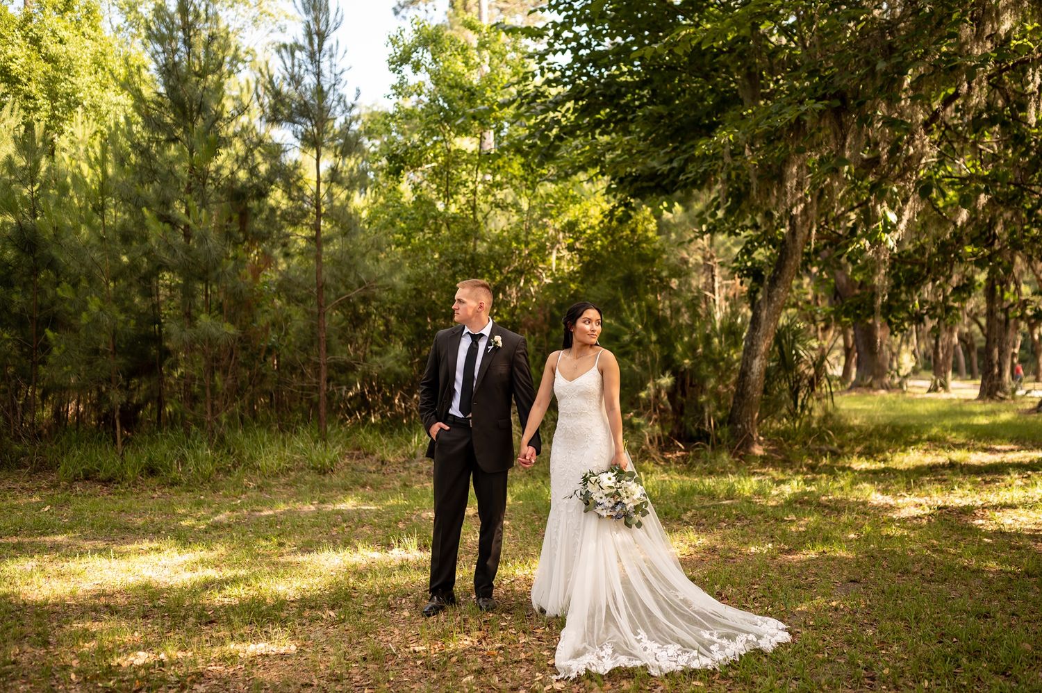 Beautiful wedding couple poses for romantic portraits in a sunlit wooded area with golden sunlight filtering through trees.