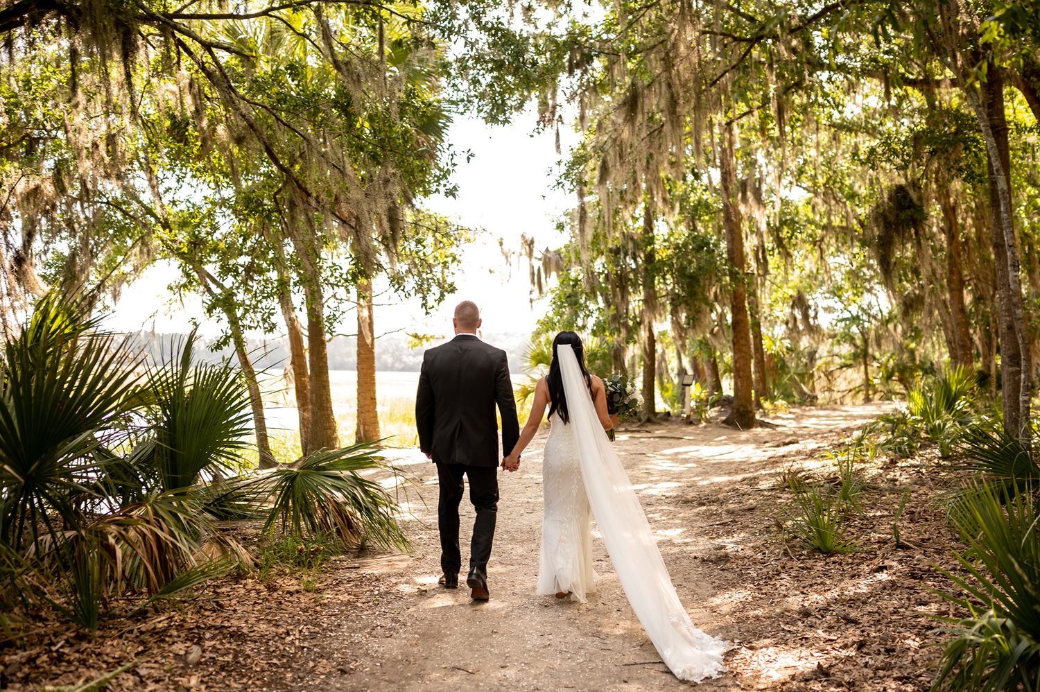 A couple walks down a sunlit path lined with moss-draped oak trees and palm fronds at sunset.