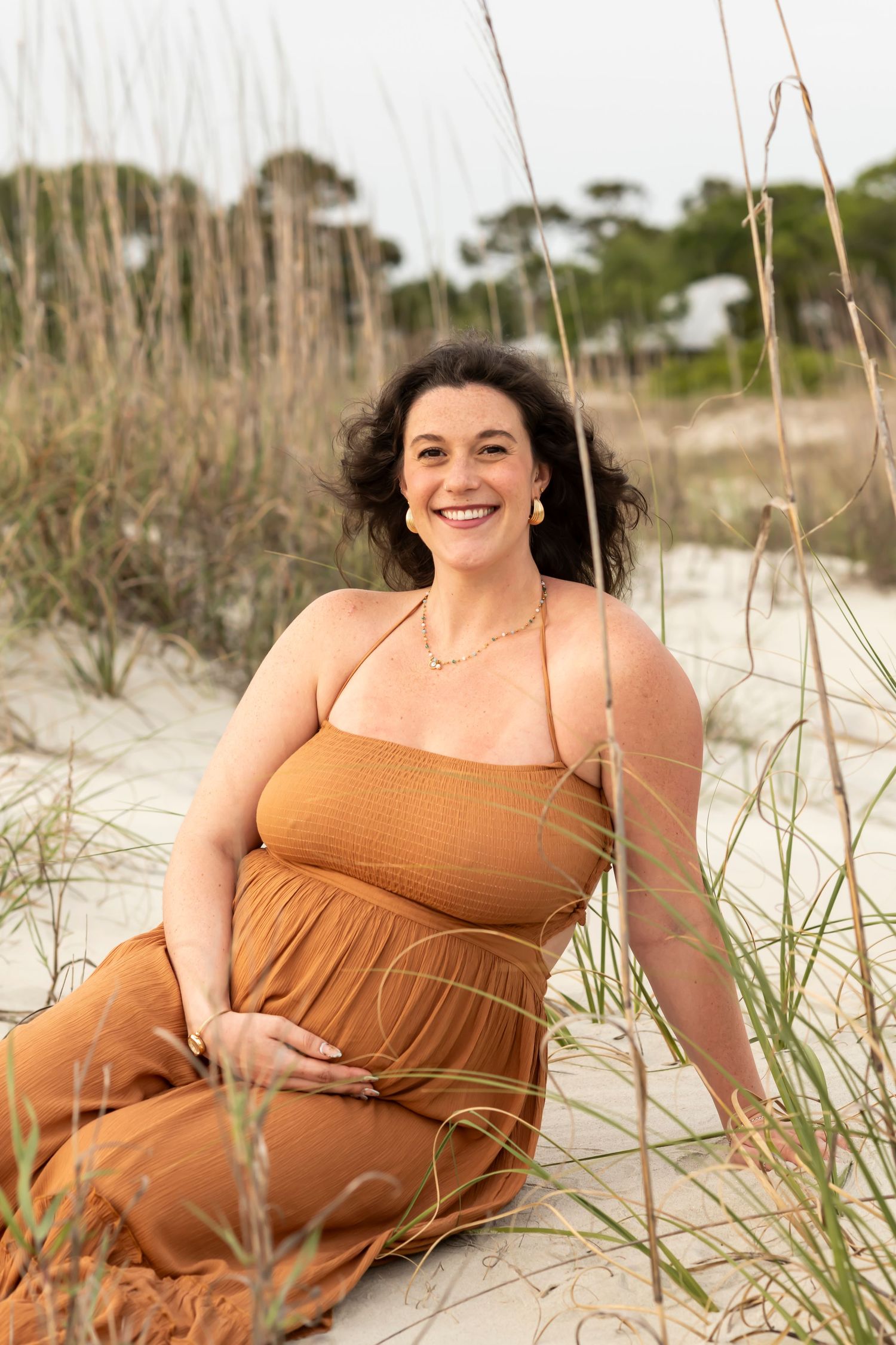 Person in rusty orange maxi dress smiles while sitting among beach dunes with sea grass on a sunny day.