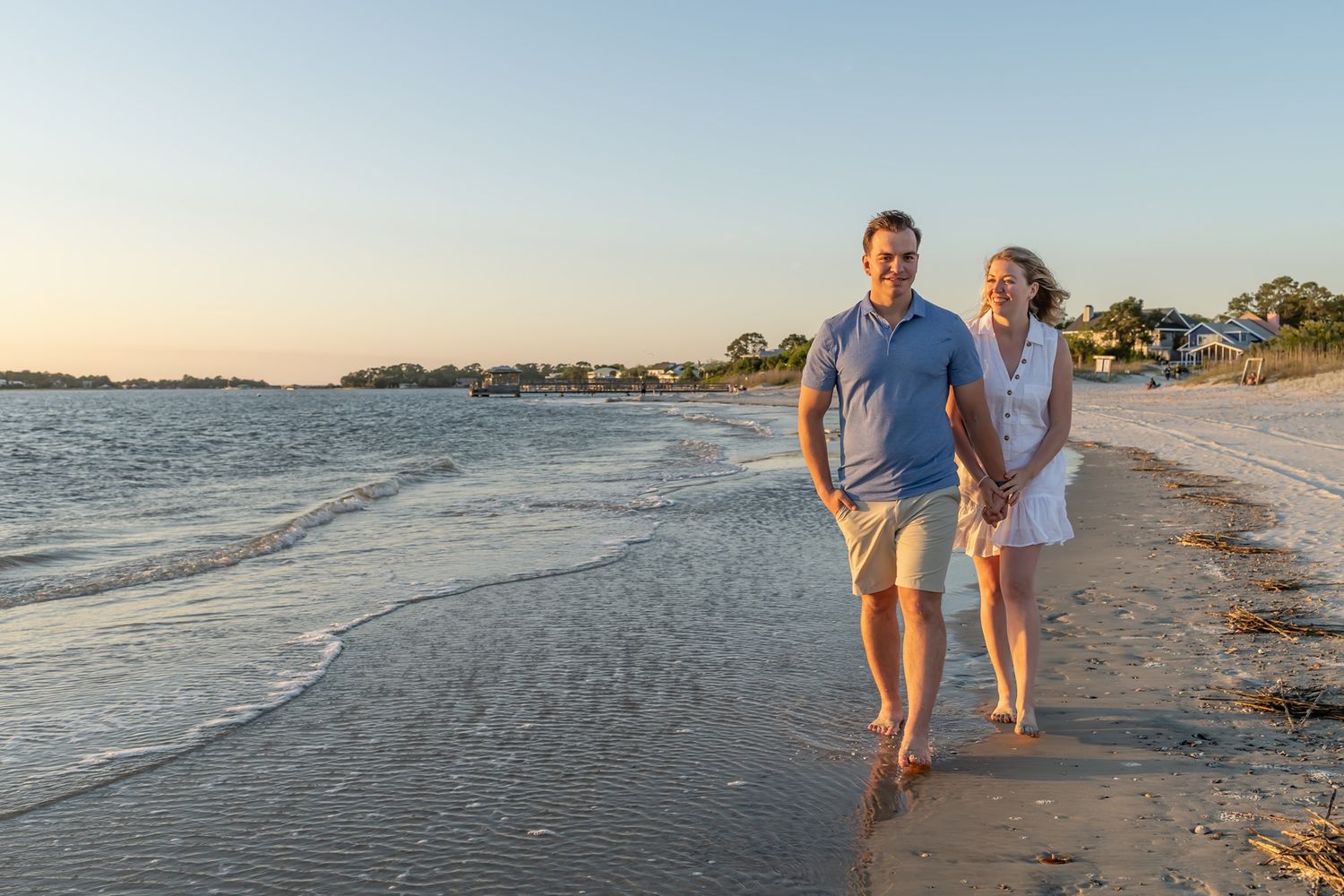 A couple walks barefoot along a sandy beach at sunset, with calm waters and coastline in the background.