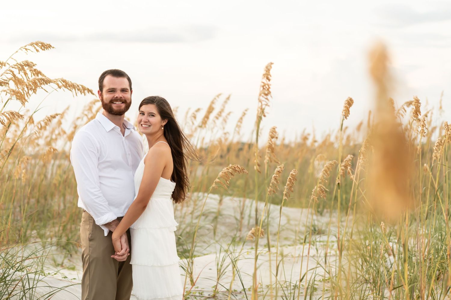 A couple stands together among tall sea oats on a sandy beach at sunset.