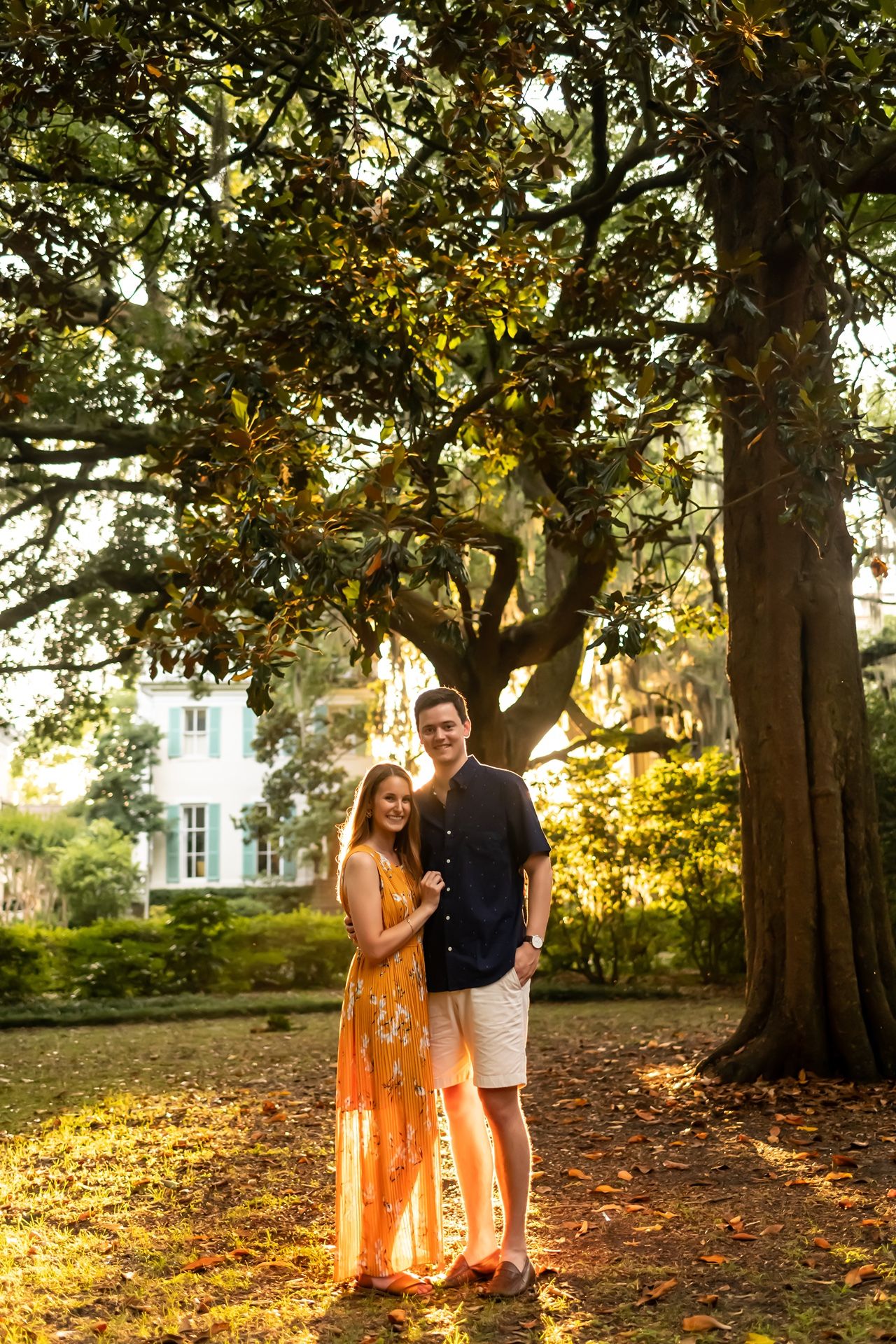 A couple embraces in a sunlit garden setting with tall trees and golden hour lighting casting a warm glow.