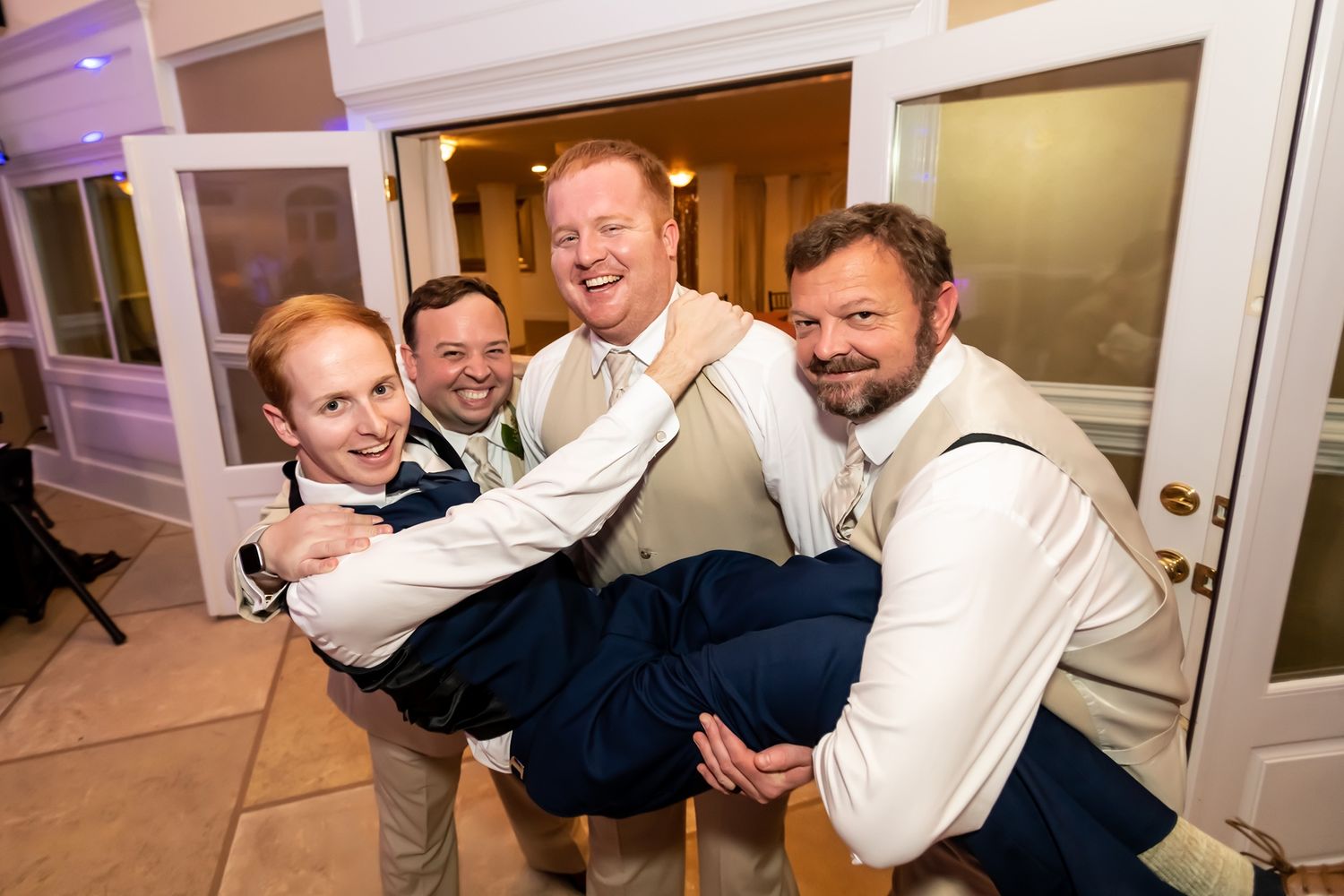 Groomsmen in white dress shirts lifting someone playfully during wedding celebrations.
