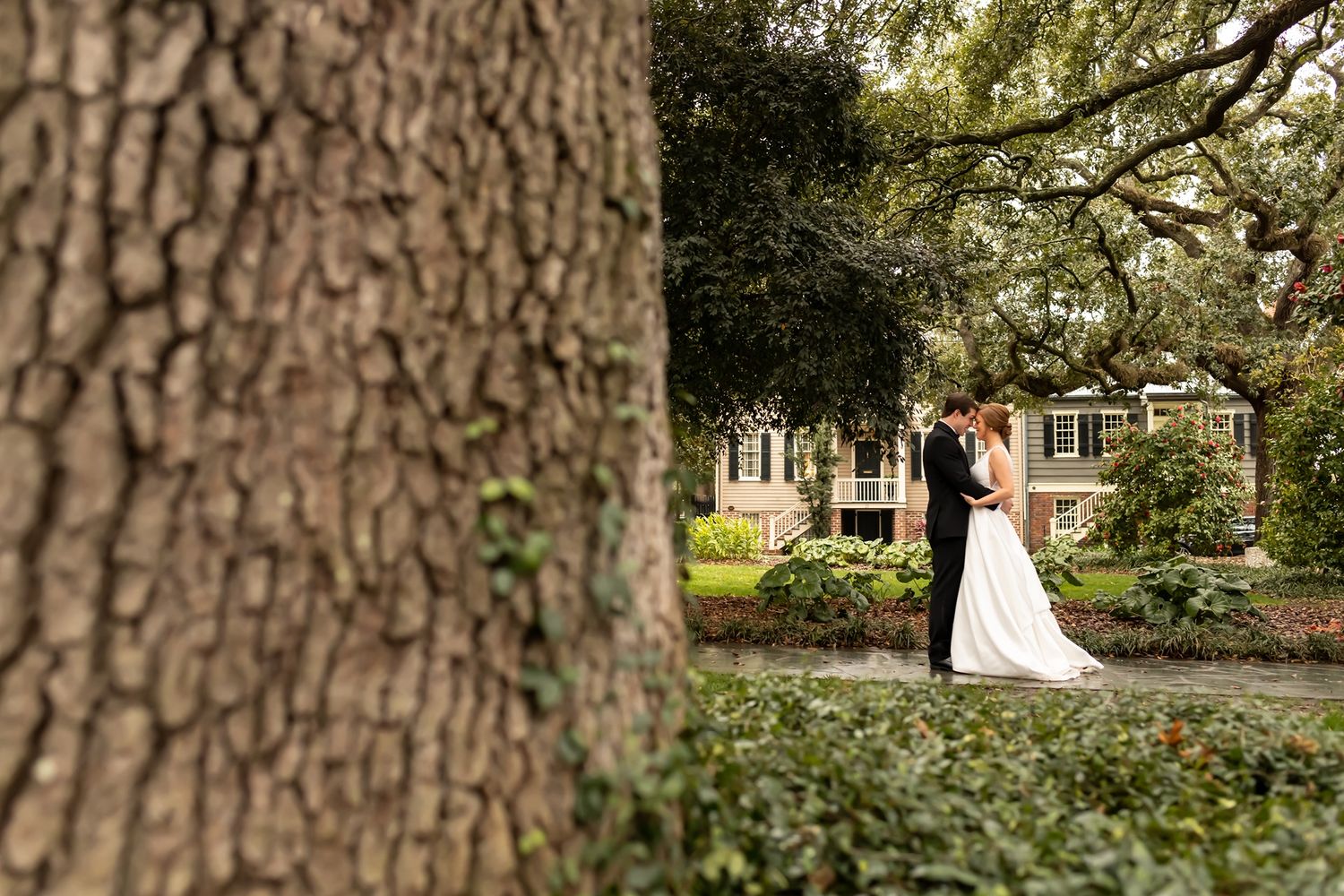 A couple shares an intimate moment together under large oak trees in a lush garden setting during their wedding celebration.