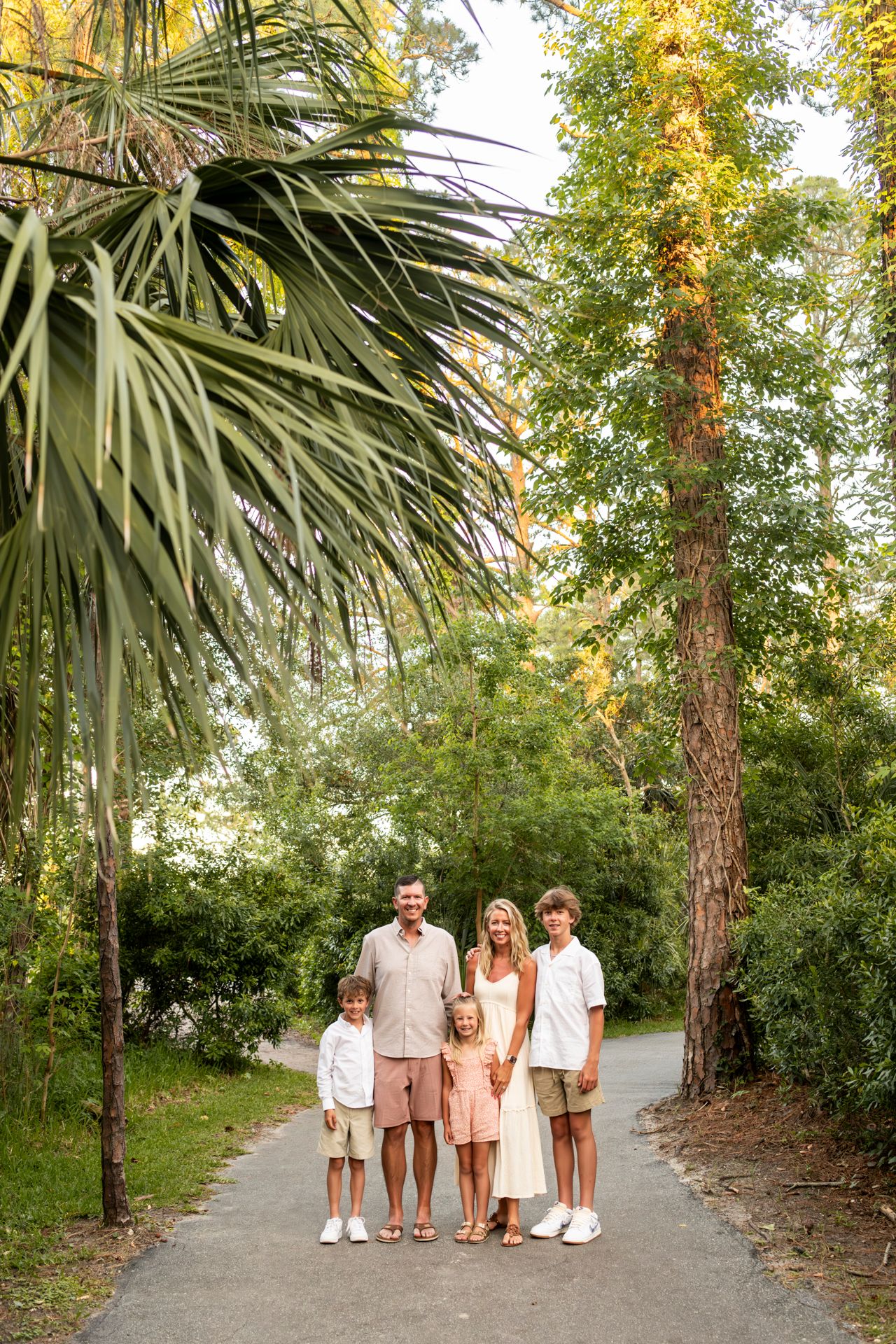 Family wearing coordinated neutral outfits stands together on a palm-lined path in a lush park setting.
