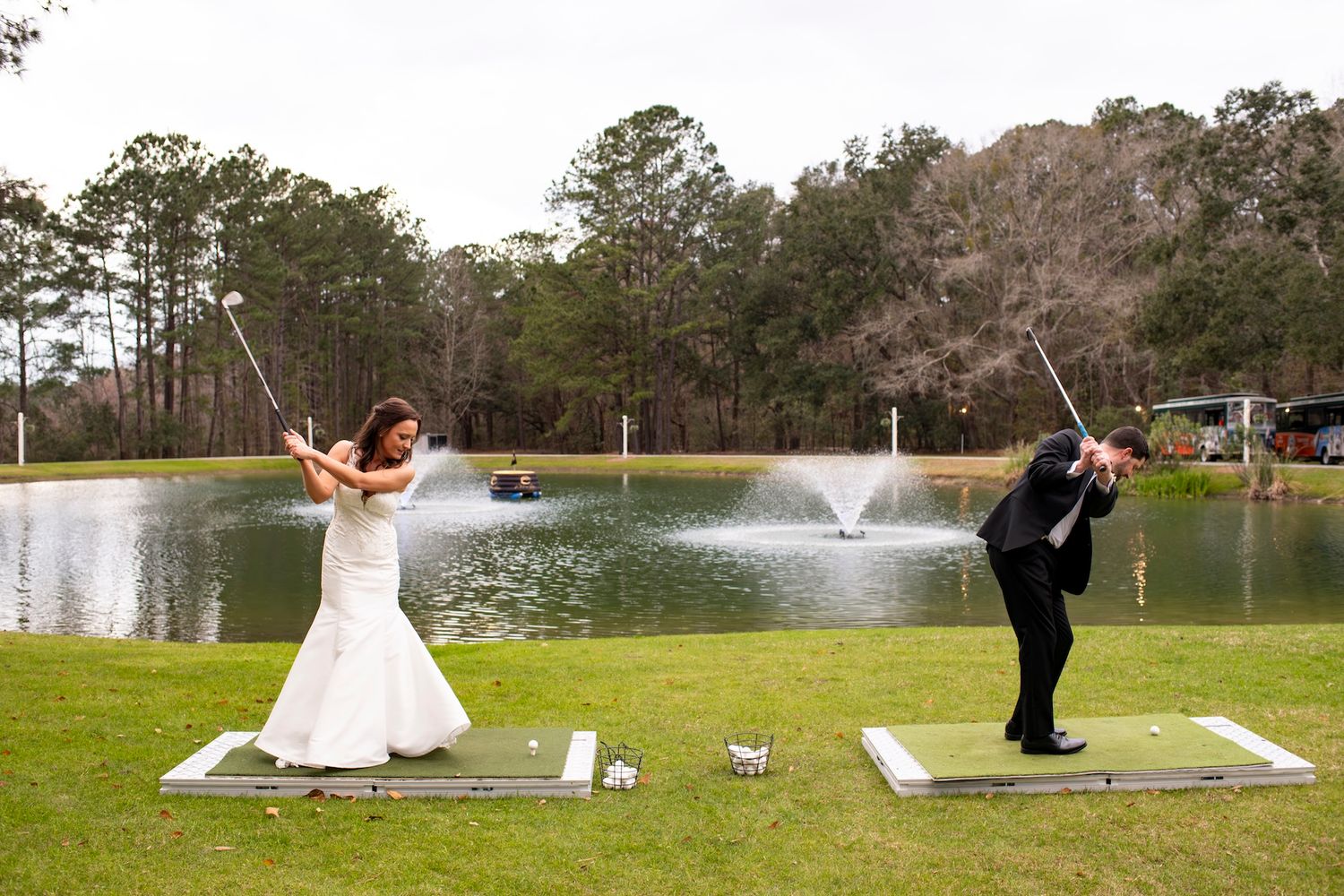 Bride and groom in wedding attire playfully swinging golf clubs near a scenic pond with fountain.