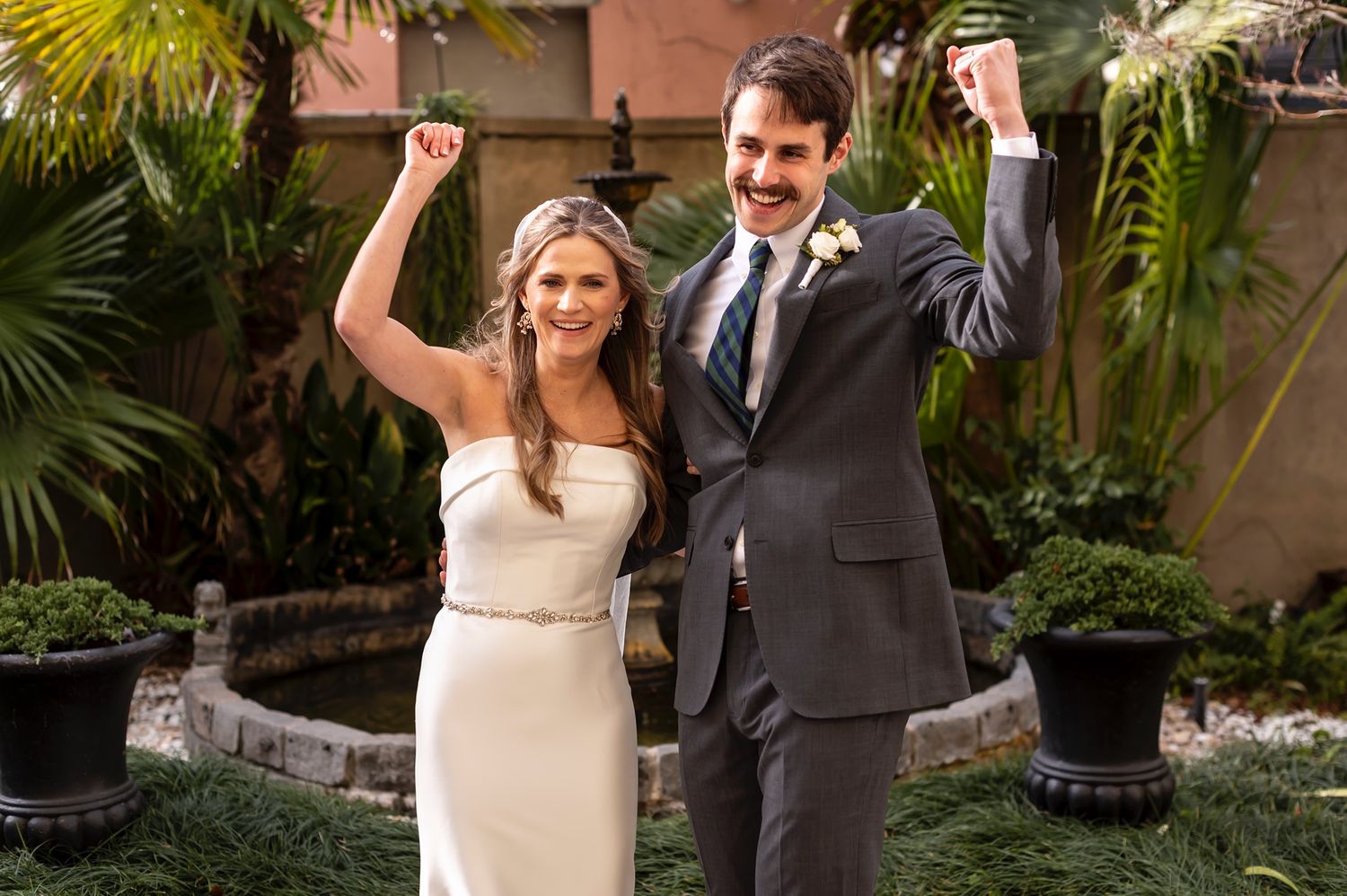 A couple in wedding attire dances and celebrates in a tropical garden setting with palm trees and greenery.