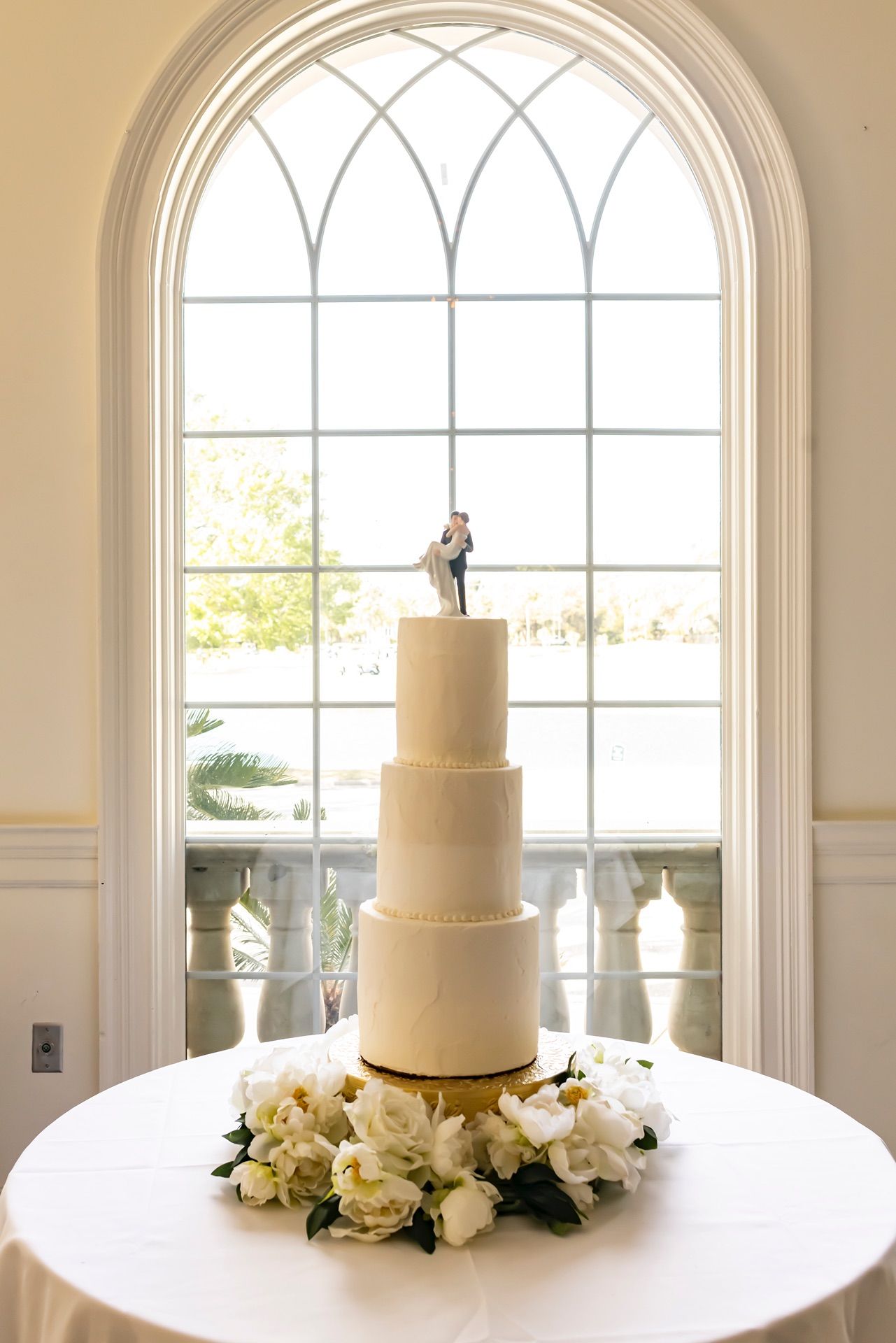 Three-tiered white wedding cake with small figurine topper displayed in front of arched window with natural light.