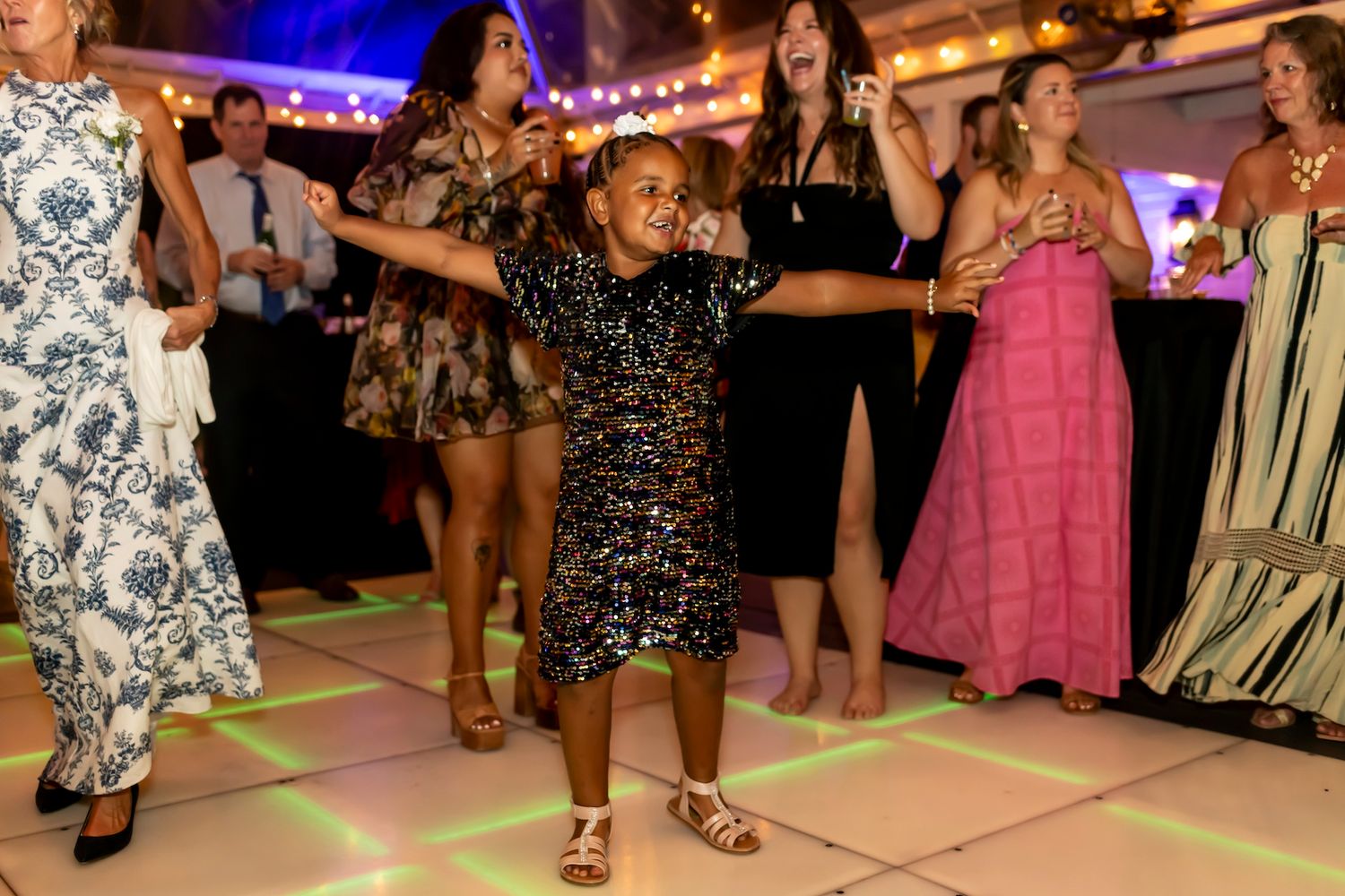 A person in a sparkly dress dances enthusiastically on a colorful light-up dance floor at a party.