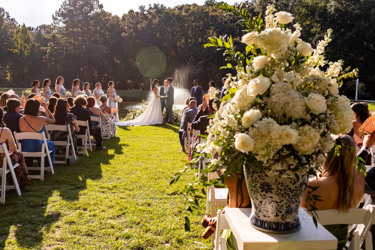 A stunning outdoor wedding ceremony with guests seated on folding chairs and an elegant white floral arrangement in the foreground.