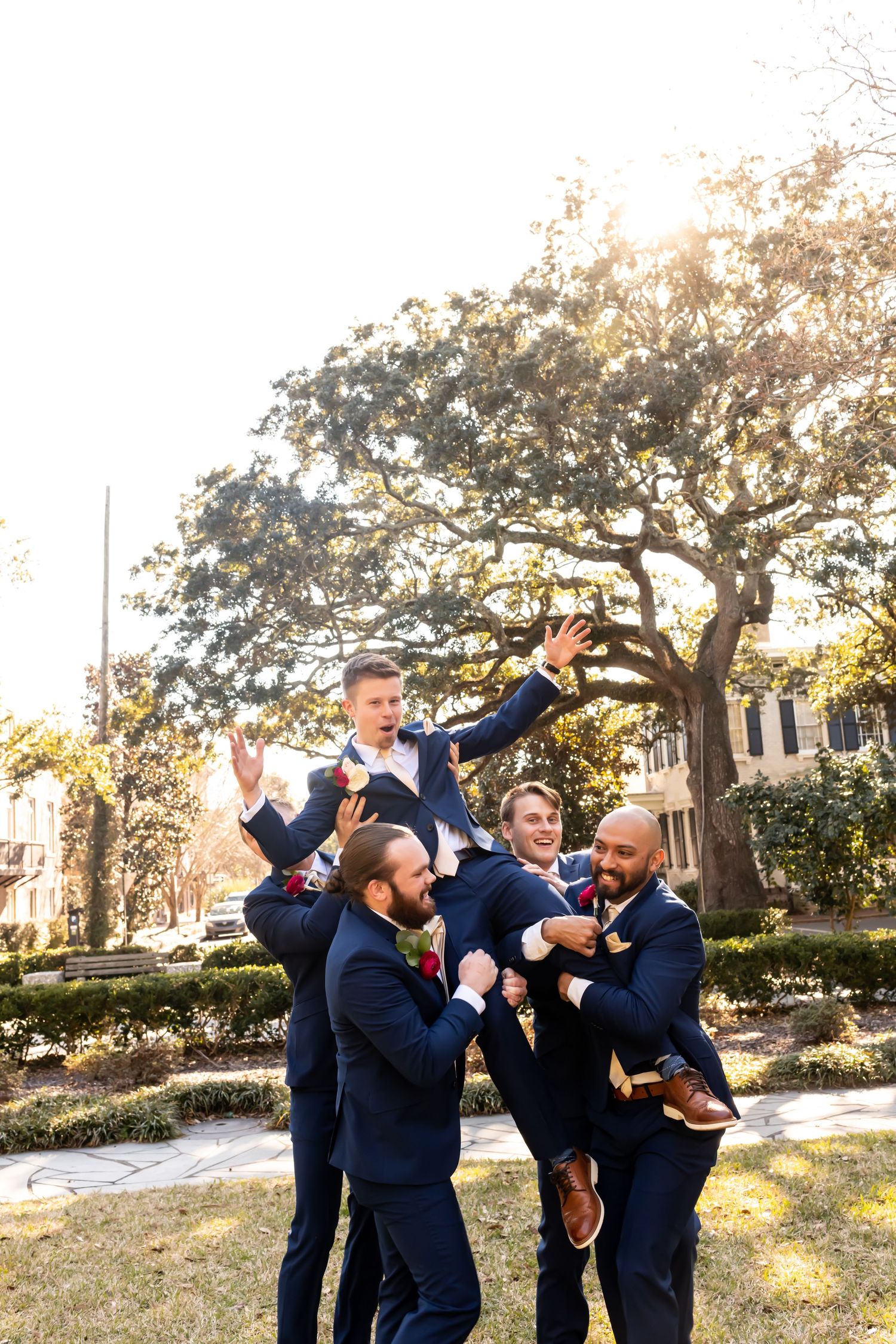 Groomsmen in navy suits celebrating joyfully outdoors under a large tree during golden hour.