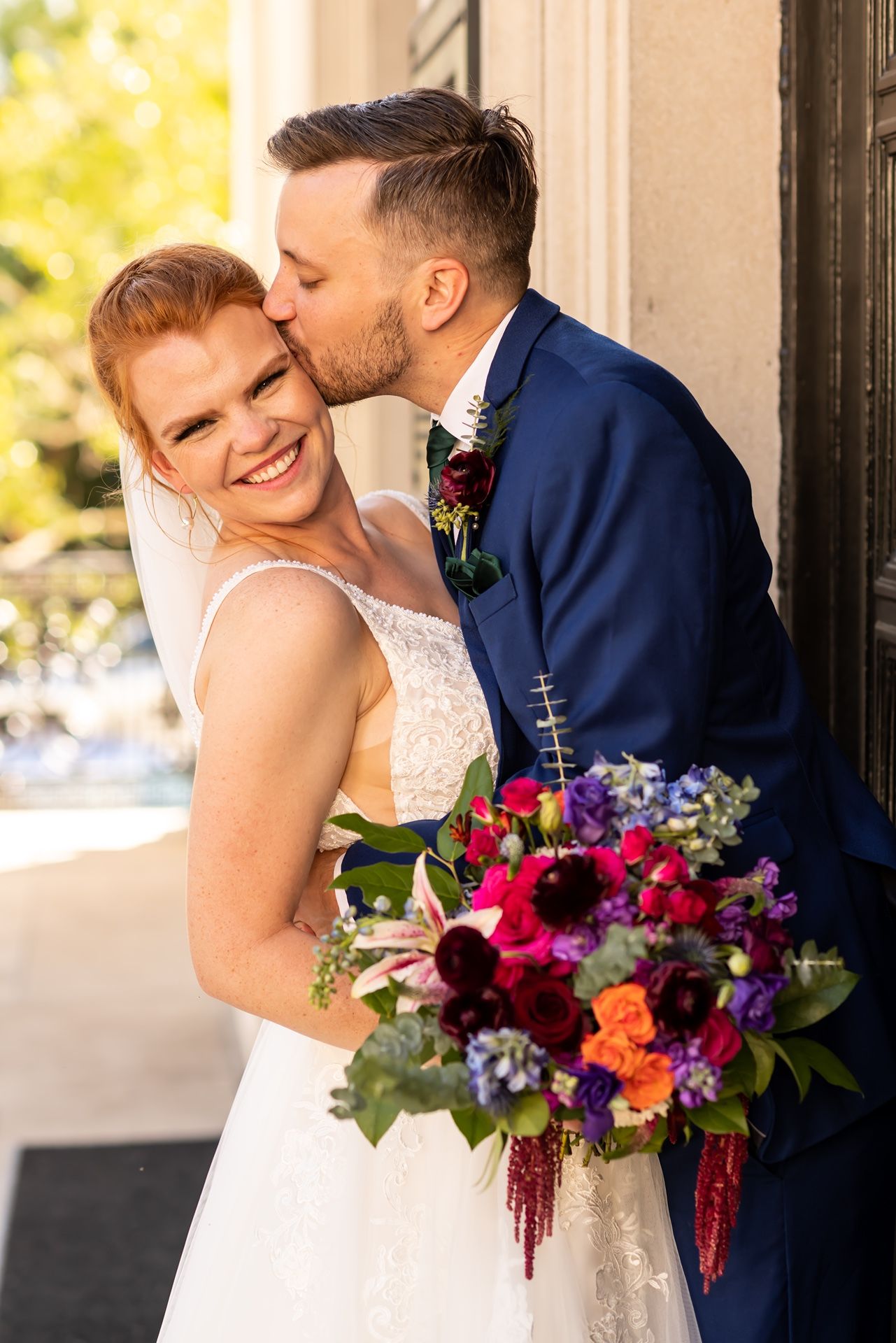A couple celebrate their wedding day as groom kisses bride while she holds vibrant multicolored bouquet in sunlit setting.