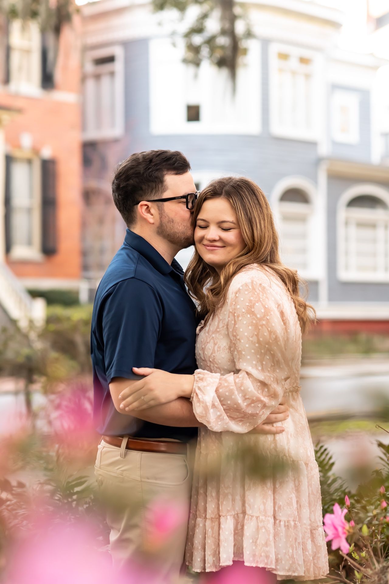 Romantic couple embracing outdoors with pink flowers and colorful buildings in the background.