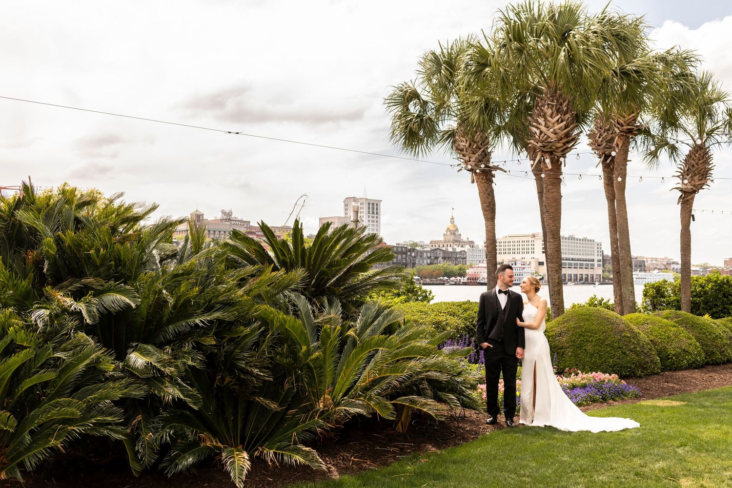 A couple stands on a lush green lawn surrounded by tropical palm trees and sago palms along a waterfront.
