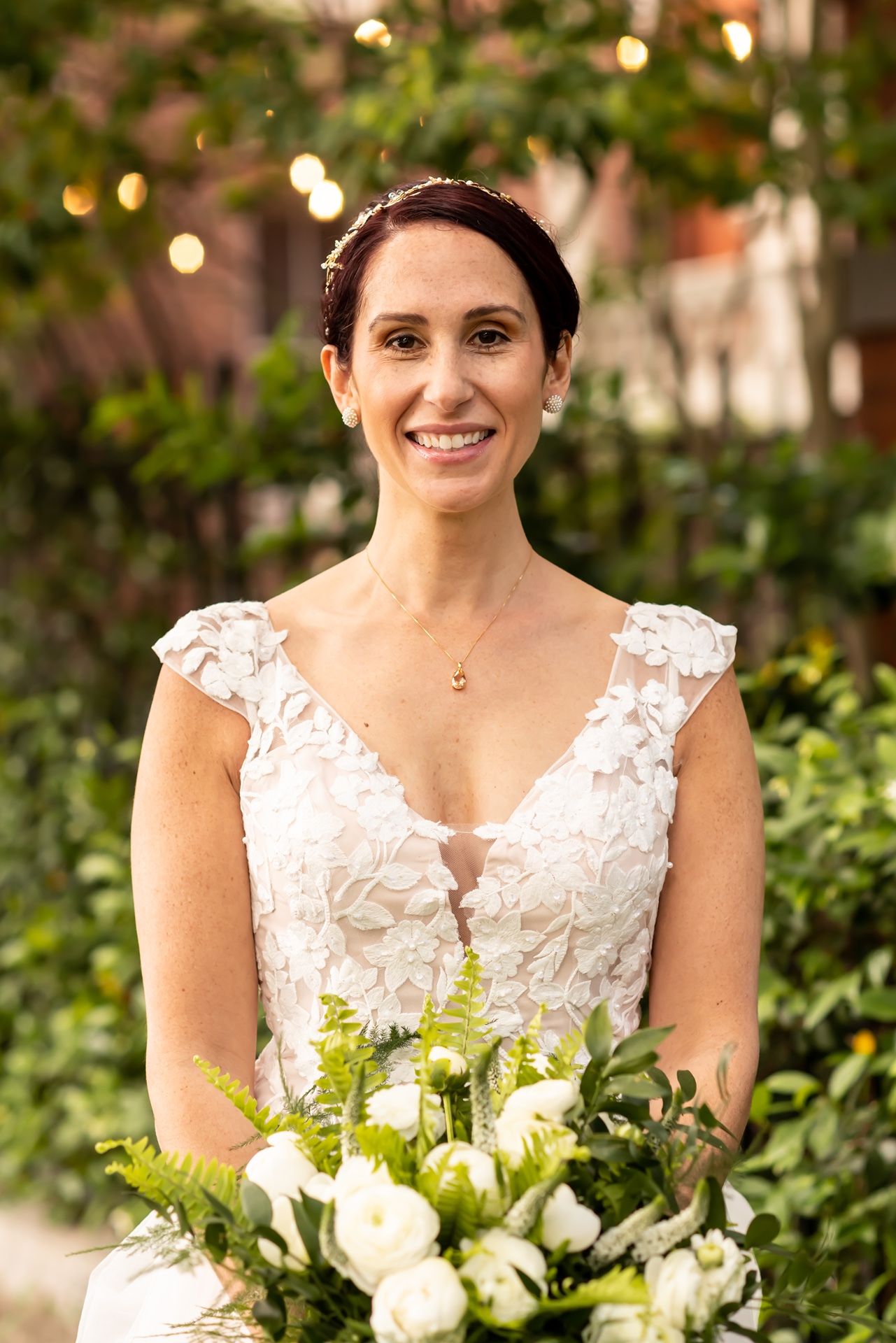 Bride wearing lace V-neck wedding dress holds white rose bouquet in garden setting with bokeh lights.