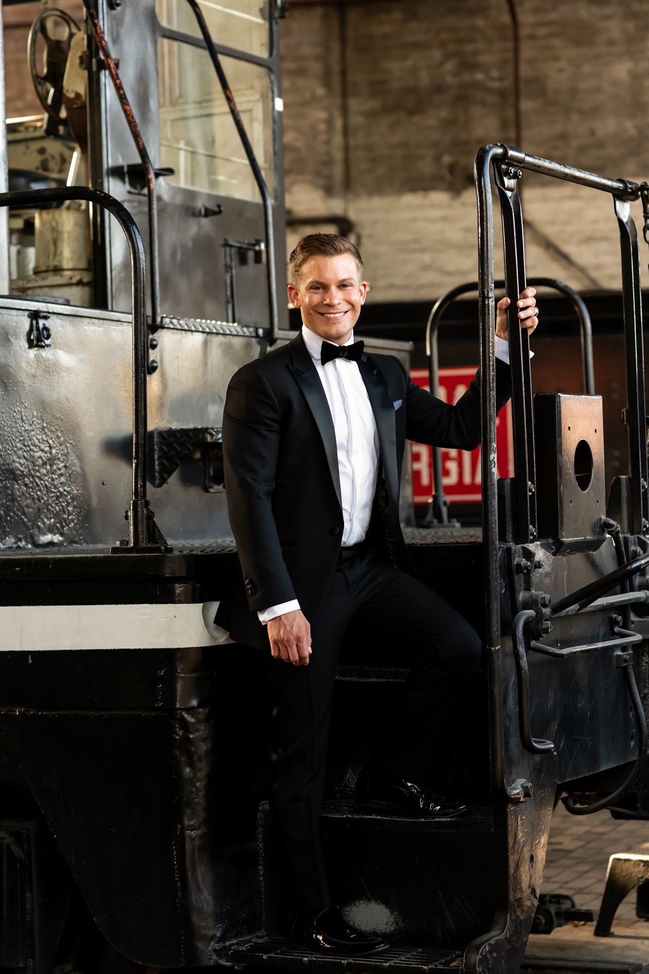 A person in a black tuxedo and bow tie stands smiling next to vintage train equipment.