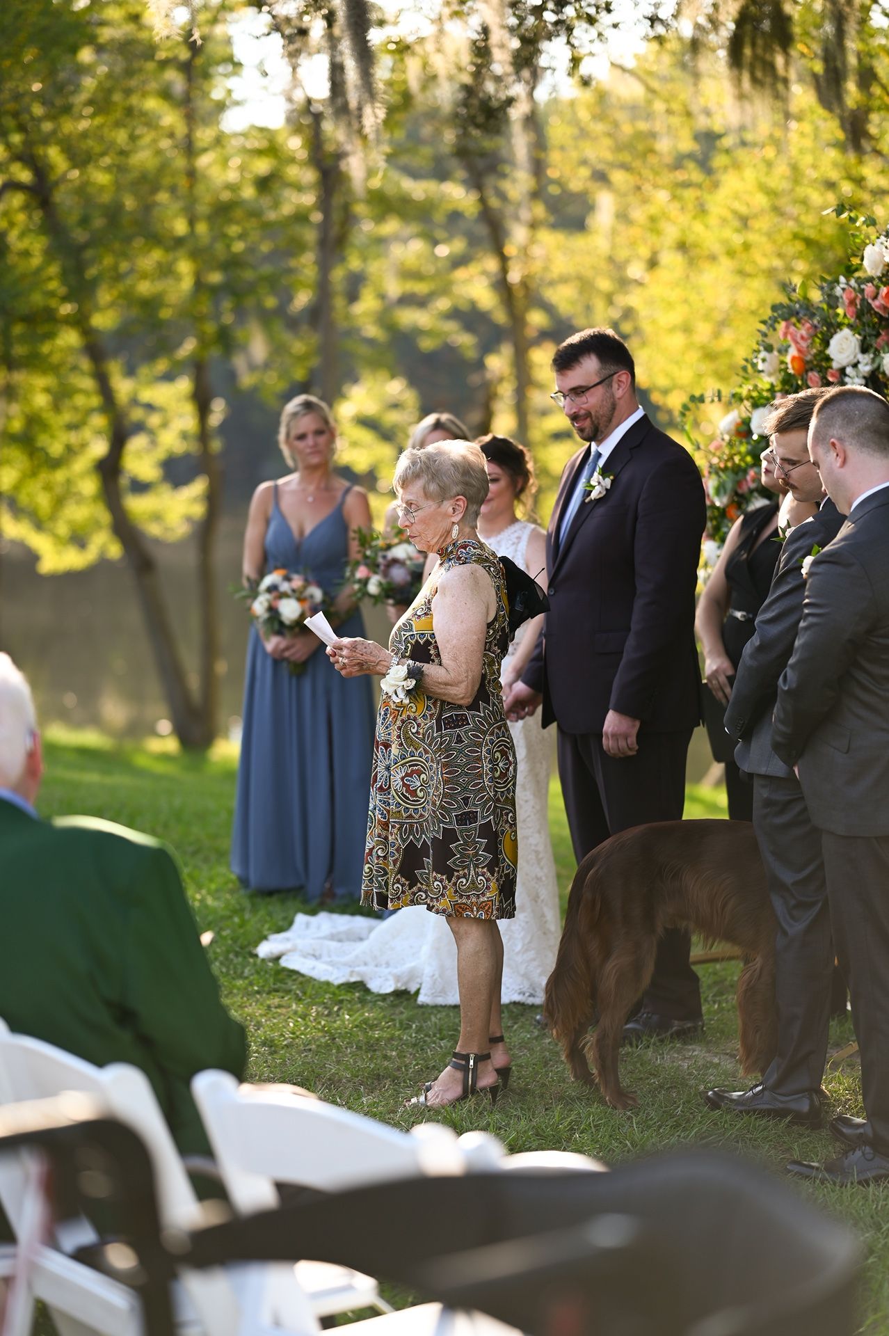 Outdoor wedding ceremony in a sunlit garden setting with attendants and floral arch arrangements.