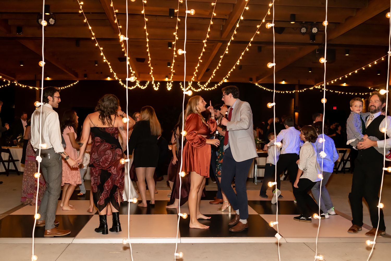 People dancing under warm string lights at an indoor reception venue with couples moving to the music on the dance floor.