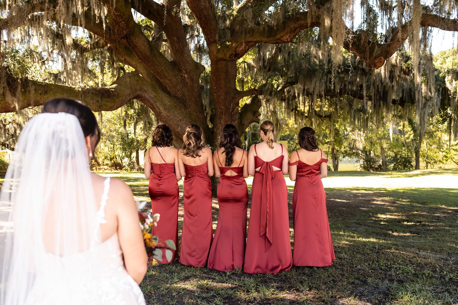 Bridesmaids in matching red gowns stand beneath a majestic oak tree draped with Spanish moss.