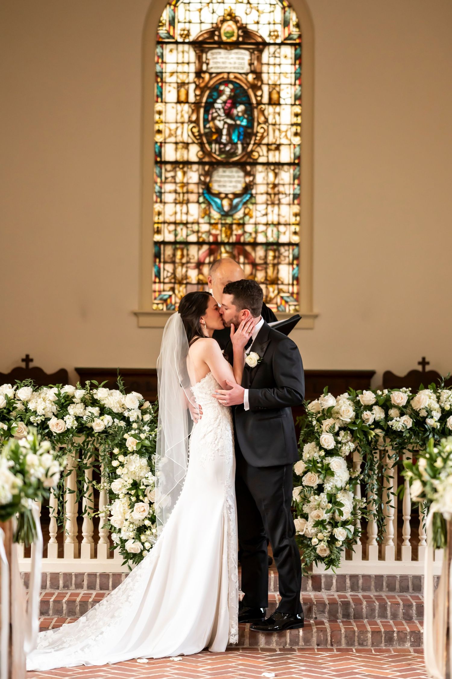 Two newlyweds share a kiss at altar with floral decor and stained glass window in church sanctuary.