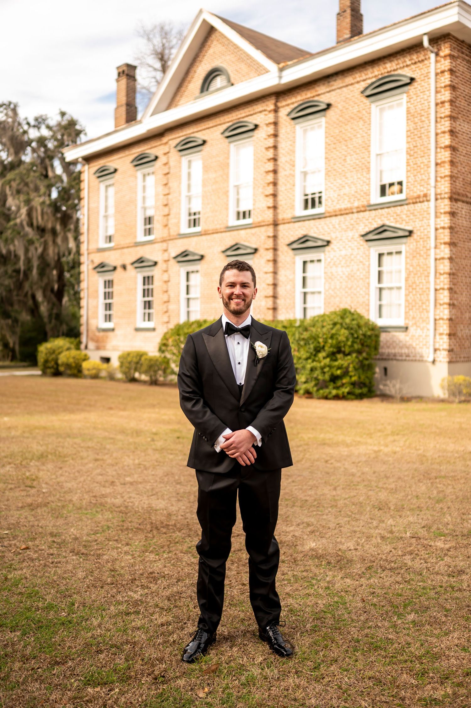 A person in formal black tuxedo and bow tie stands smiling in front of a grand historic brick mansion on a grassy lawn.