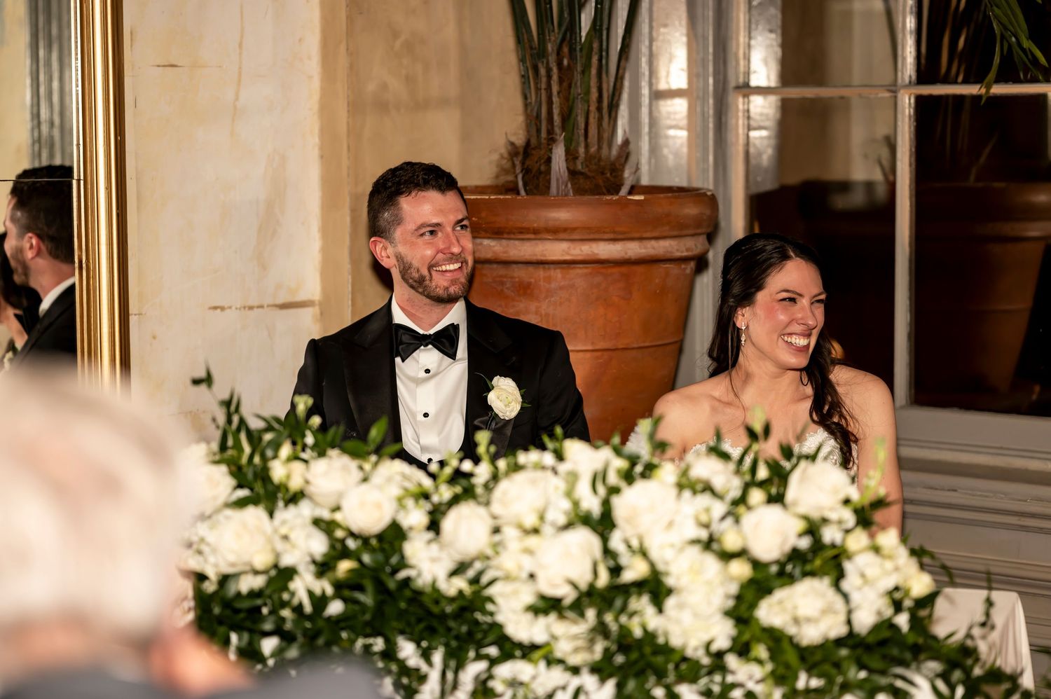 Newlyweds smile and laugh together at their reception table decorated with elegant white floral centerpieces.