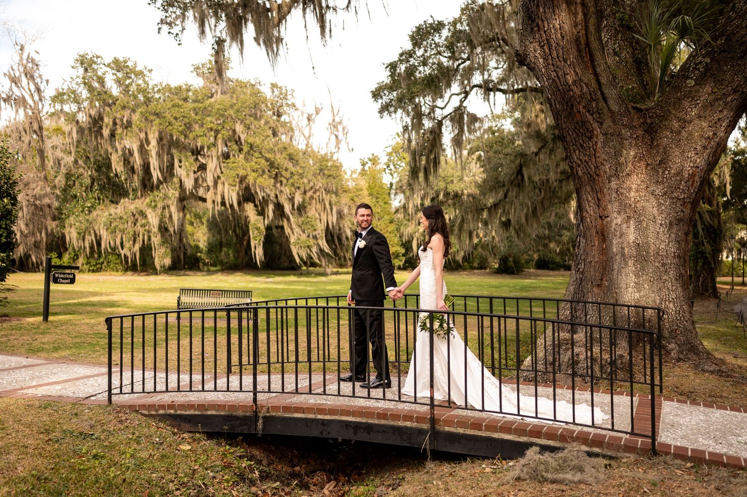 A couple poses on a rustic wooden bridge surrounded by Spanish moss-draped trees at an outdoor venue.